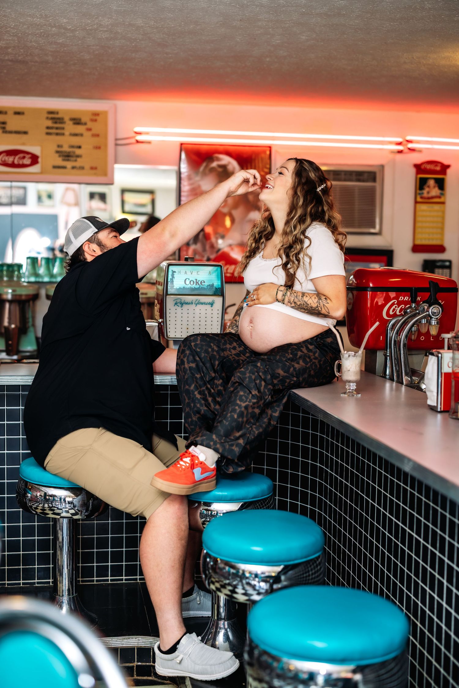 A man feeds a pregnant woman at a retro diner counter with teal stools and Coca-Cola decor.