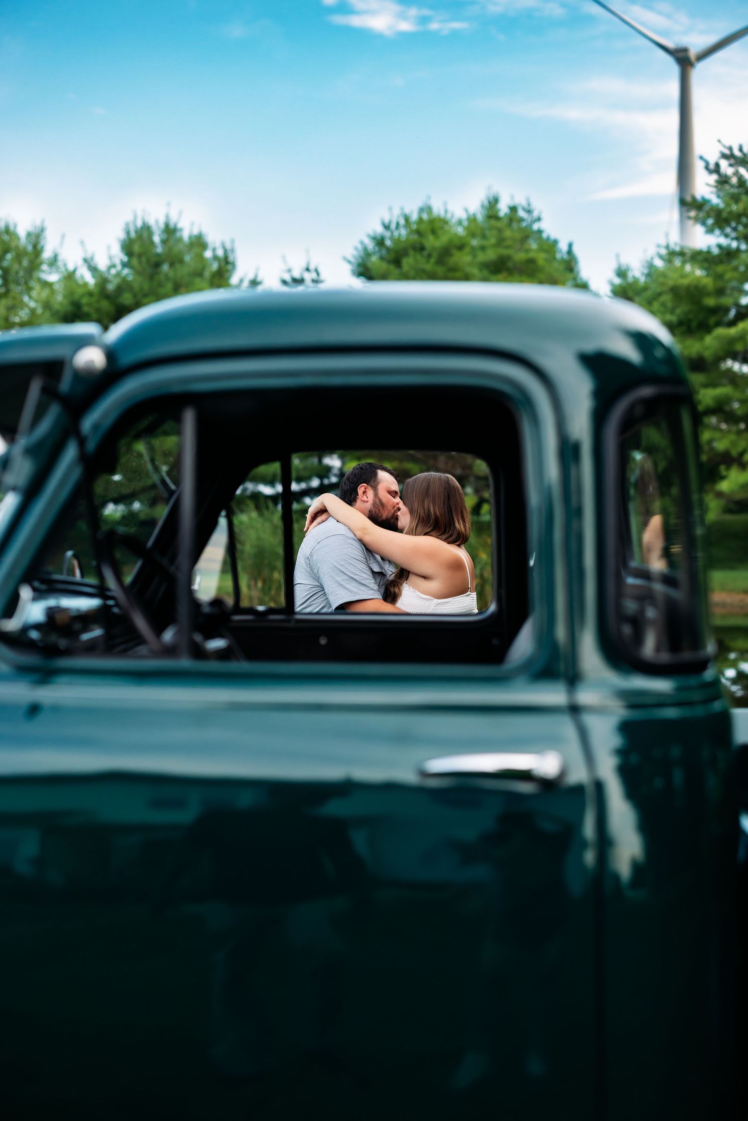 A vintage green truck's side mirror and window frame a romantic moment against a blue summer sky with trees.