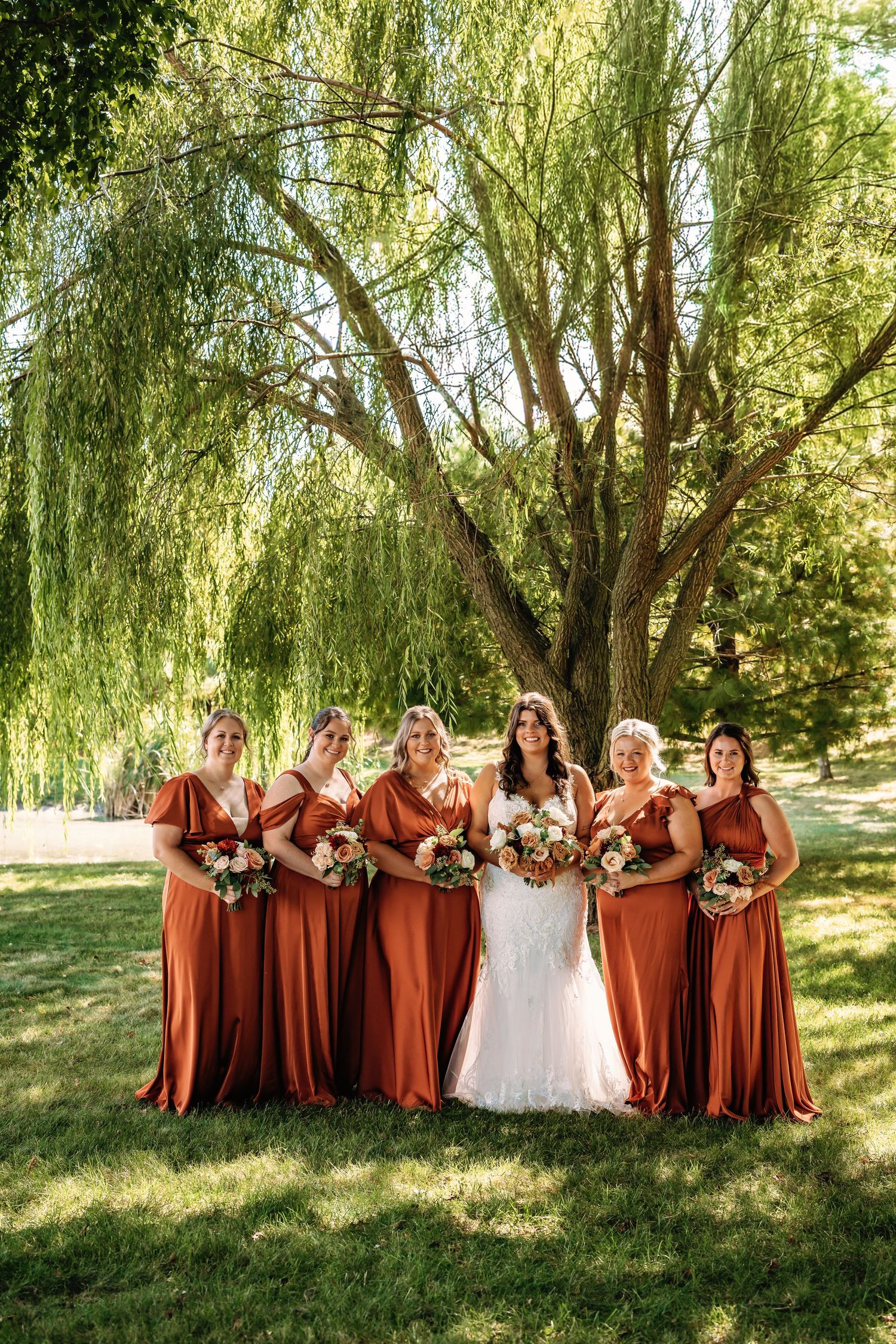 A bridal party in rust-colored dresses poses together under weeping willow trees on a sunny afternoon.