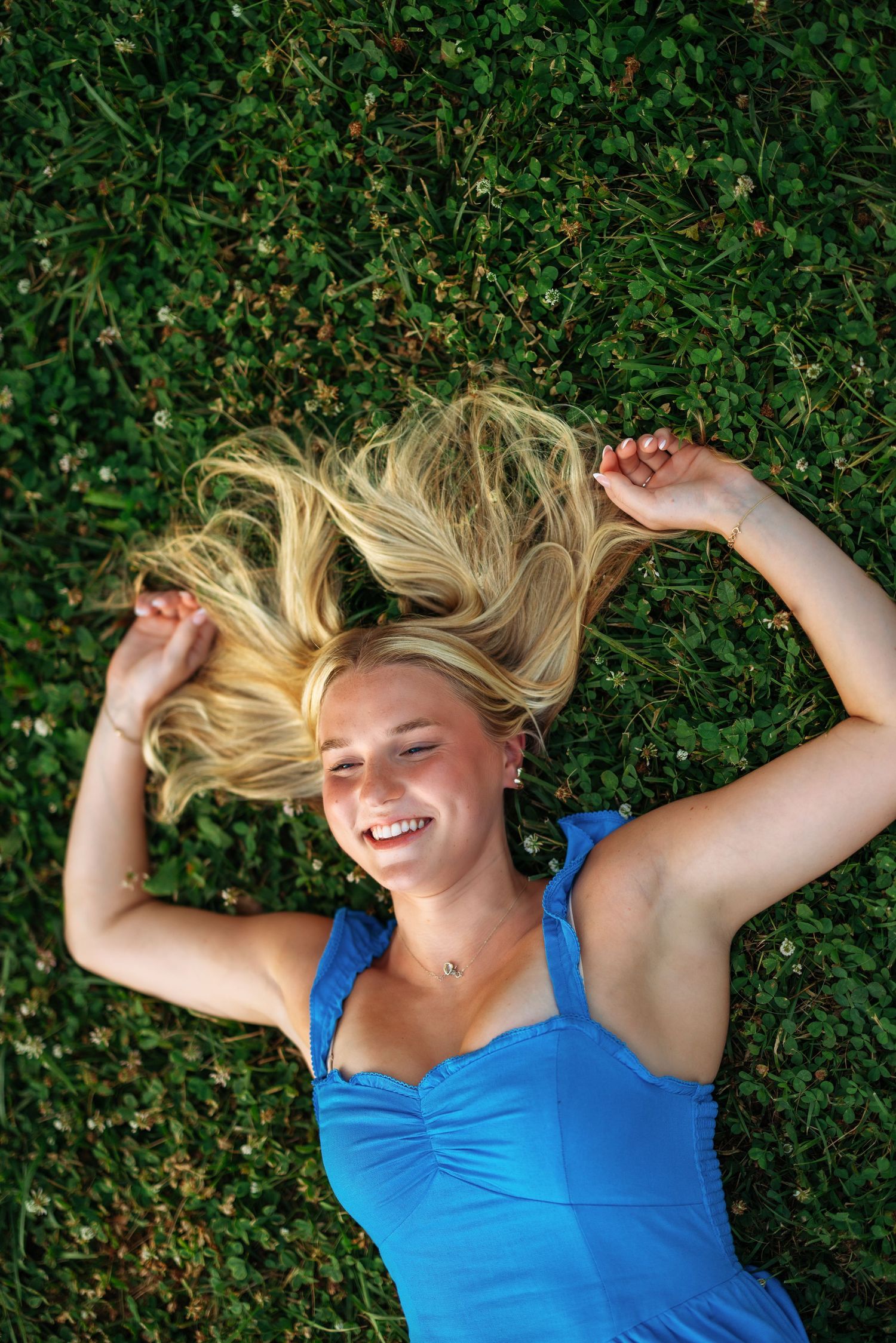 Smiling blonde woman in blue dress lying on green grass with hair spread out, looking up joyfully.