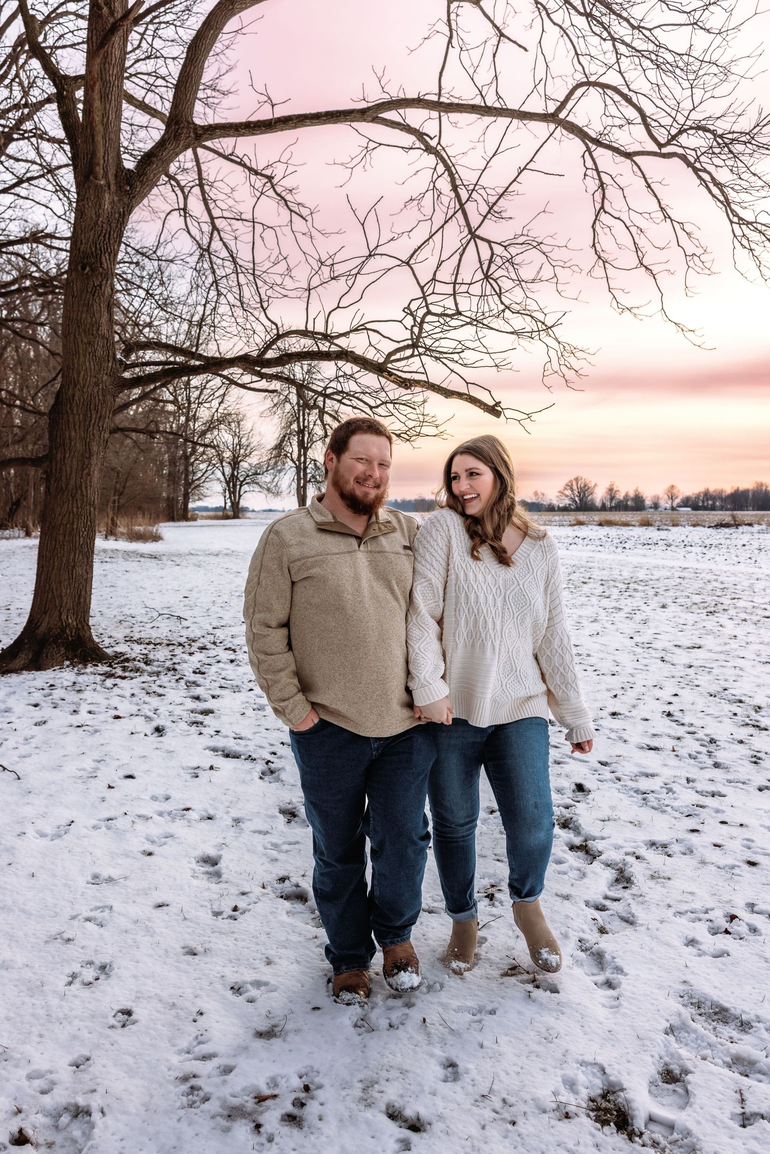 A couple shares tender moments in a snowy winter landscape with bare trees at sunset.