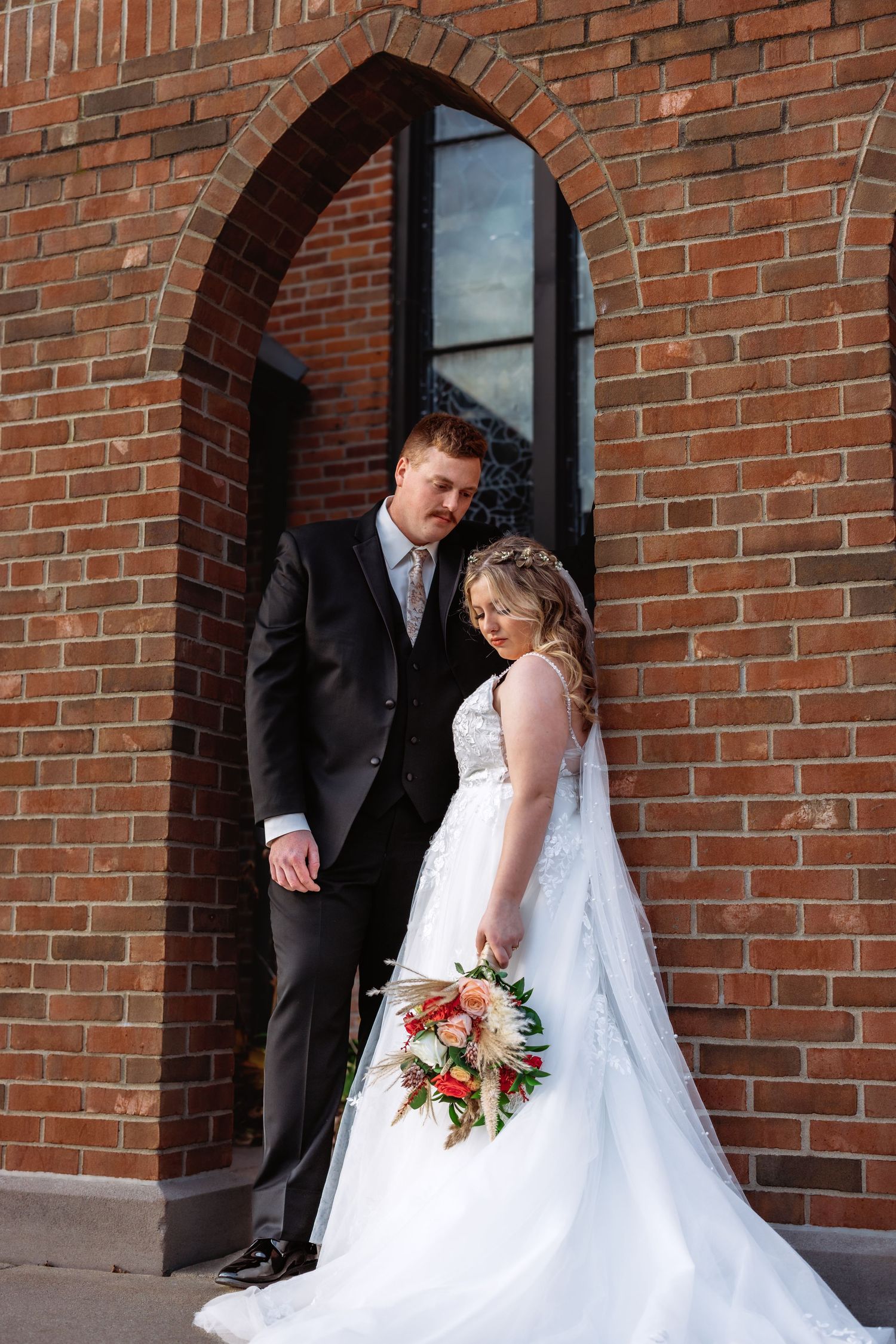 A wedding couple poses in an arched brick doorway, with the bride holding a pink and white floral bouquet.