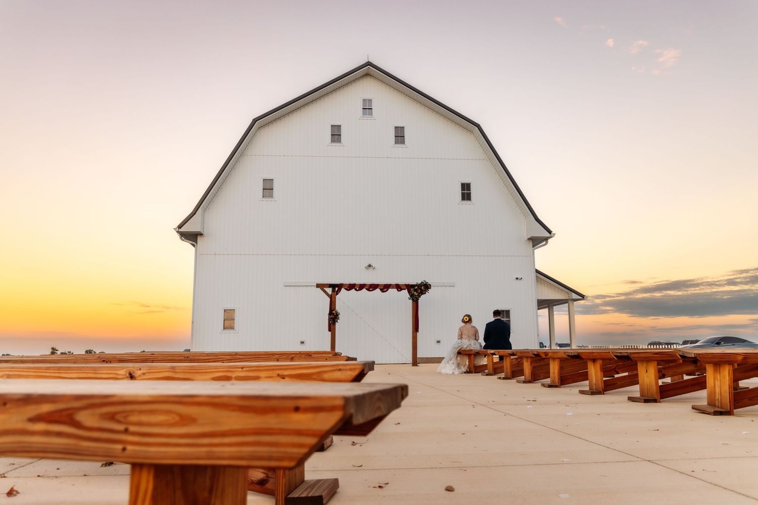 White barn wedding venue with wooden benches at sunset.