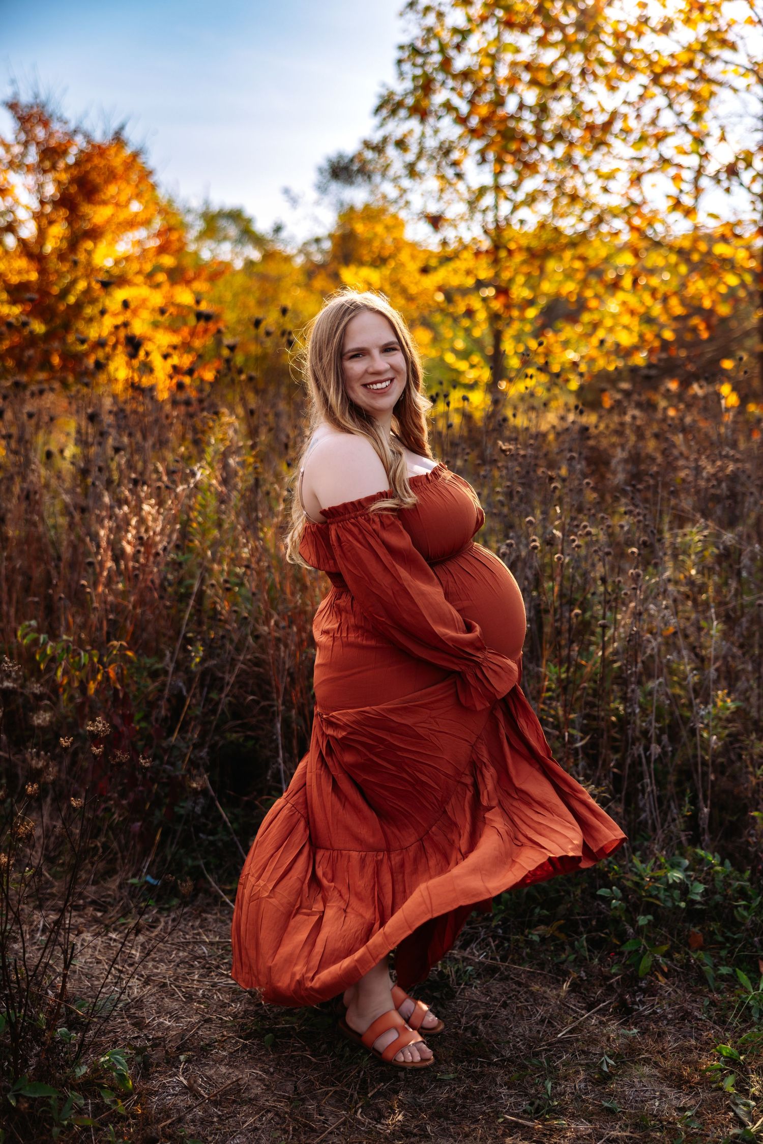 A figure in a rust-colored dress twirls playfully in a golden autumn field at sunset with fall foliage in the background.