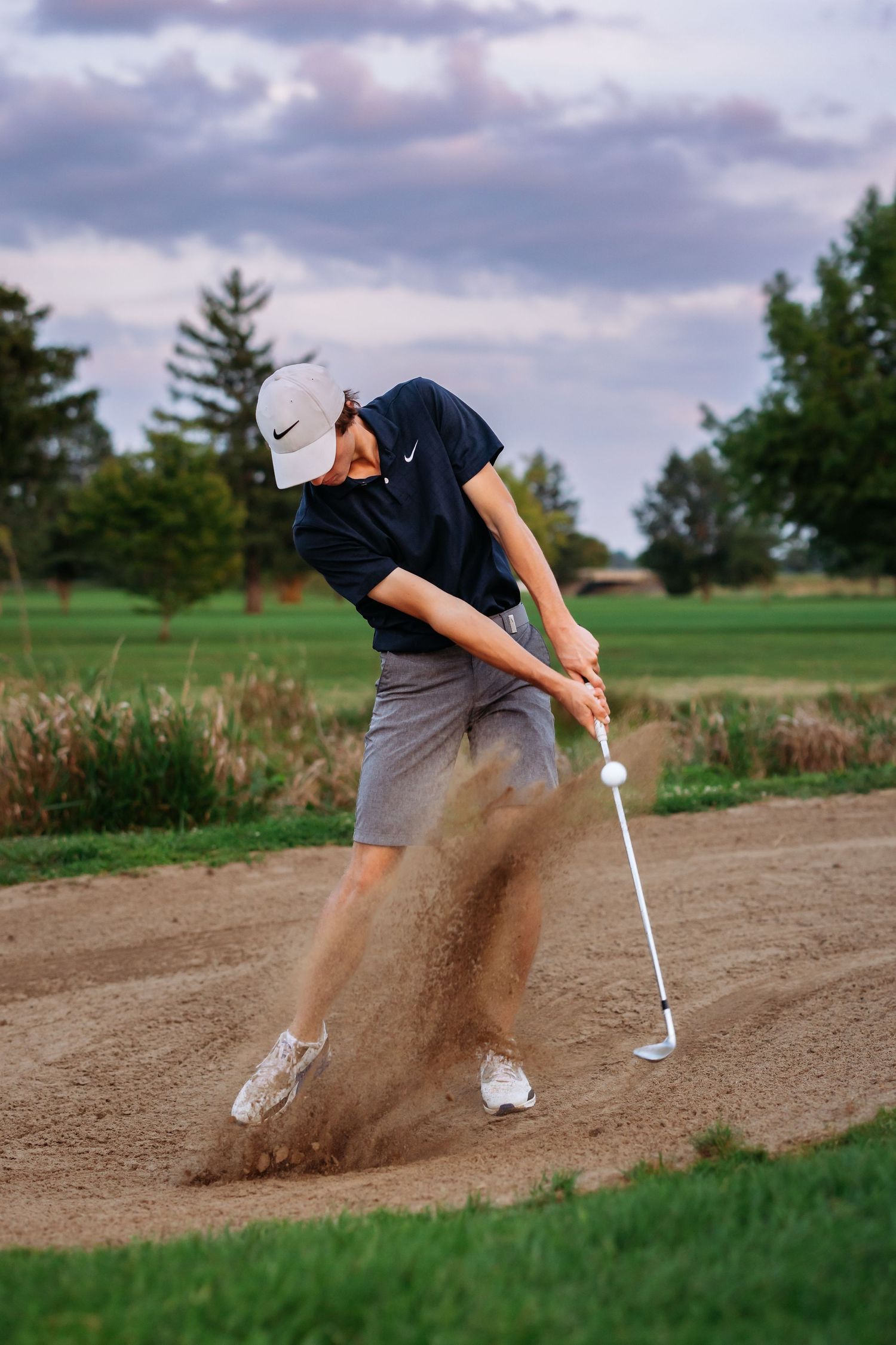 Golfer in navy shirt hitting ball from sand trap creating dramatic dust cloud.