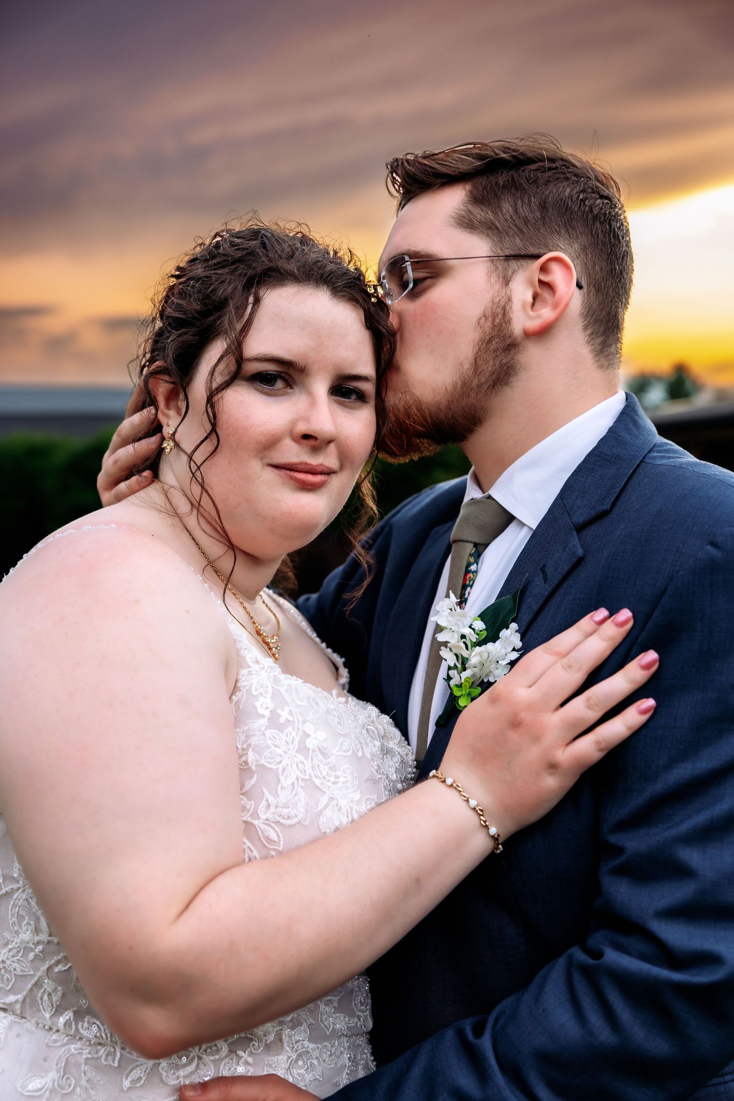 A groom in a navy suit kisses his bride in a white lace dress during a romantic sunset wedding portrait.