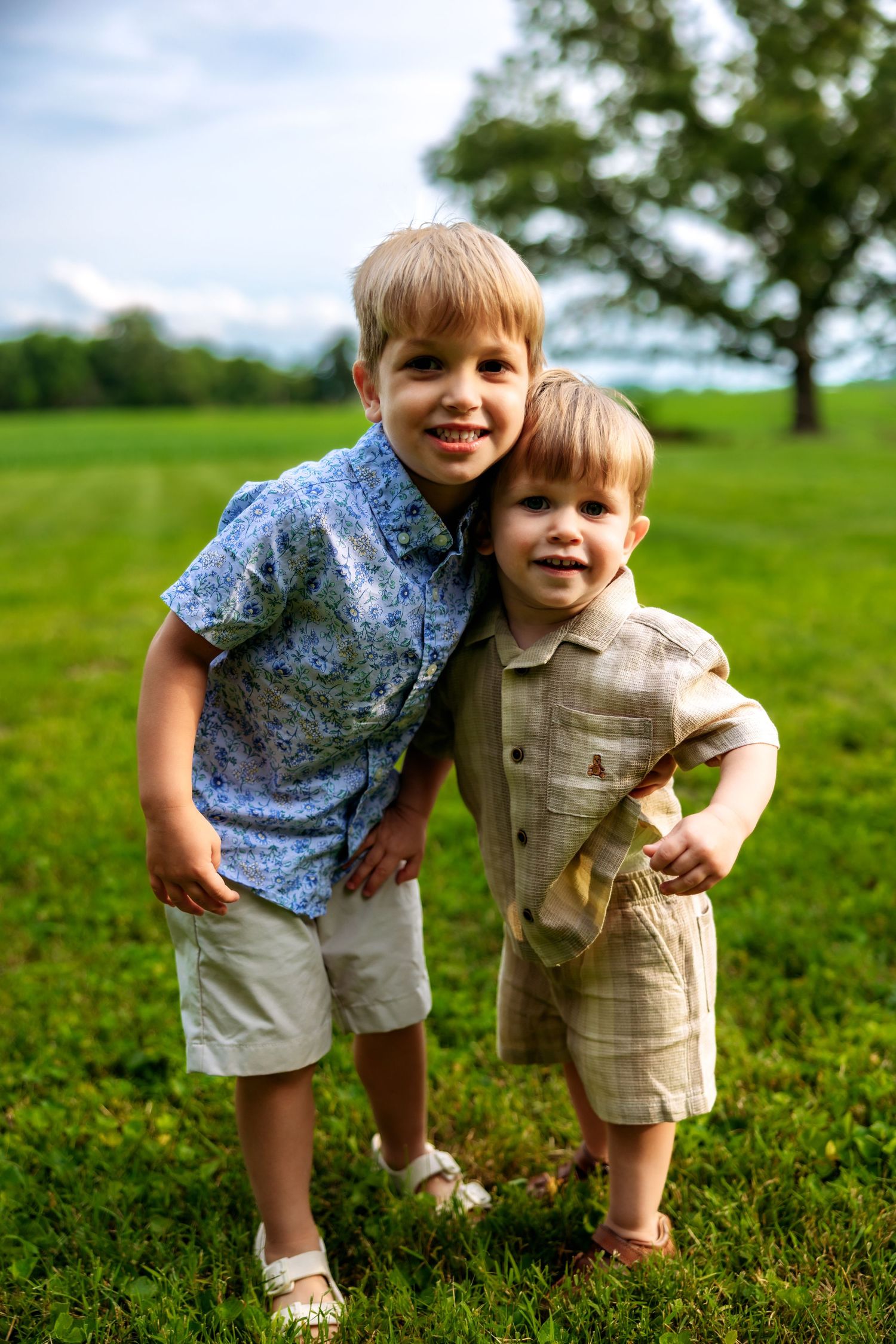 Two young boys smiling and hugging outdoors on green grass, wearing casual summer outfits on a sunny day.
