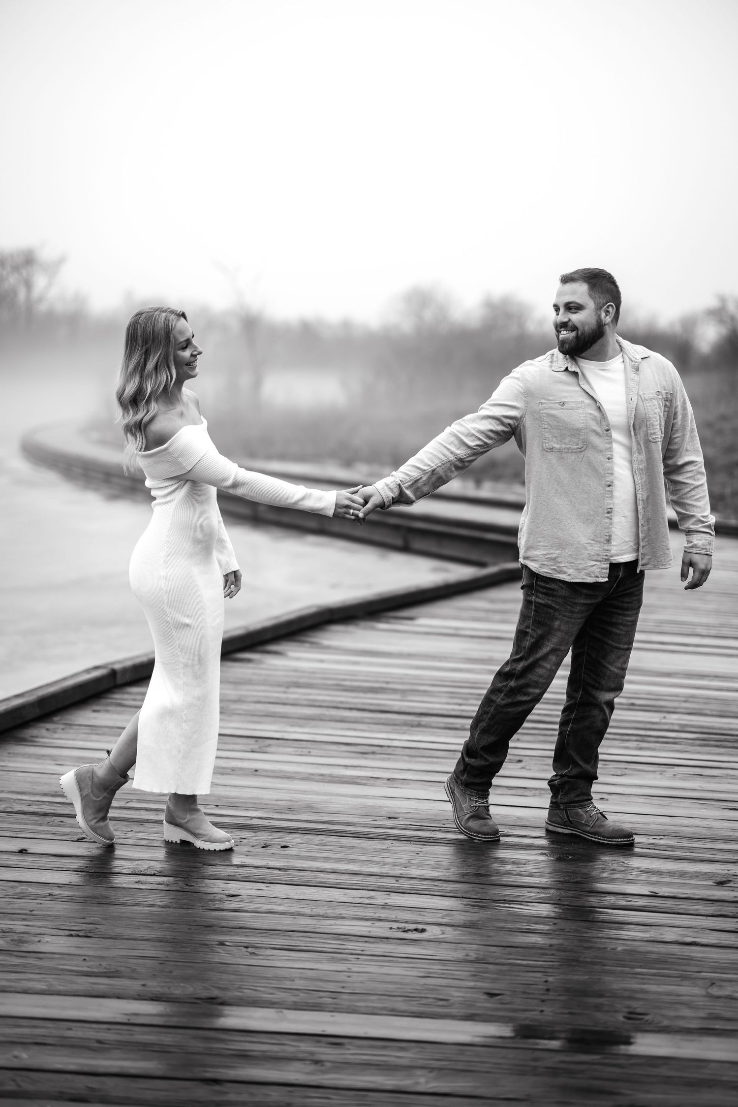 Couple holding hands playfully on wooden boardwalk in black and white photograph.
