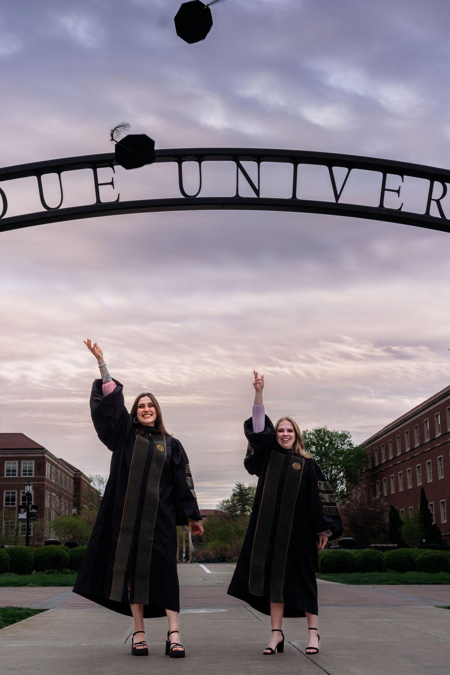 Two graduates in black robes tossing caps under university entrance sign at sunset.