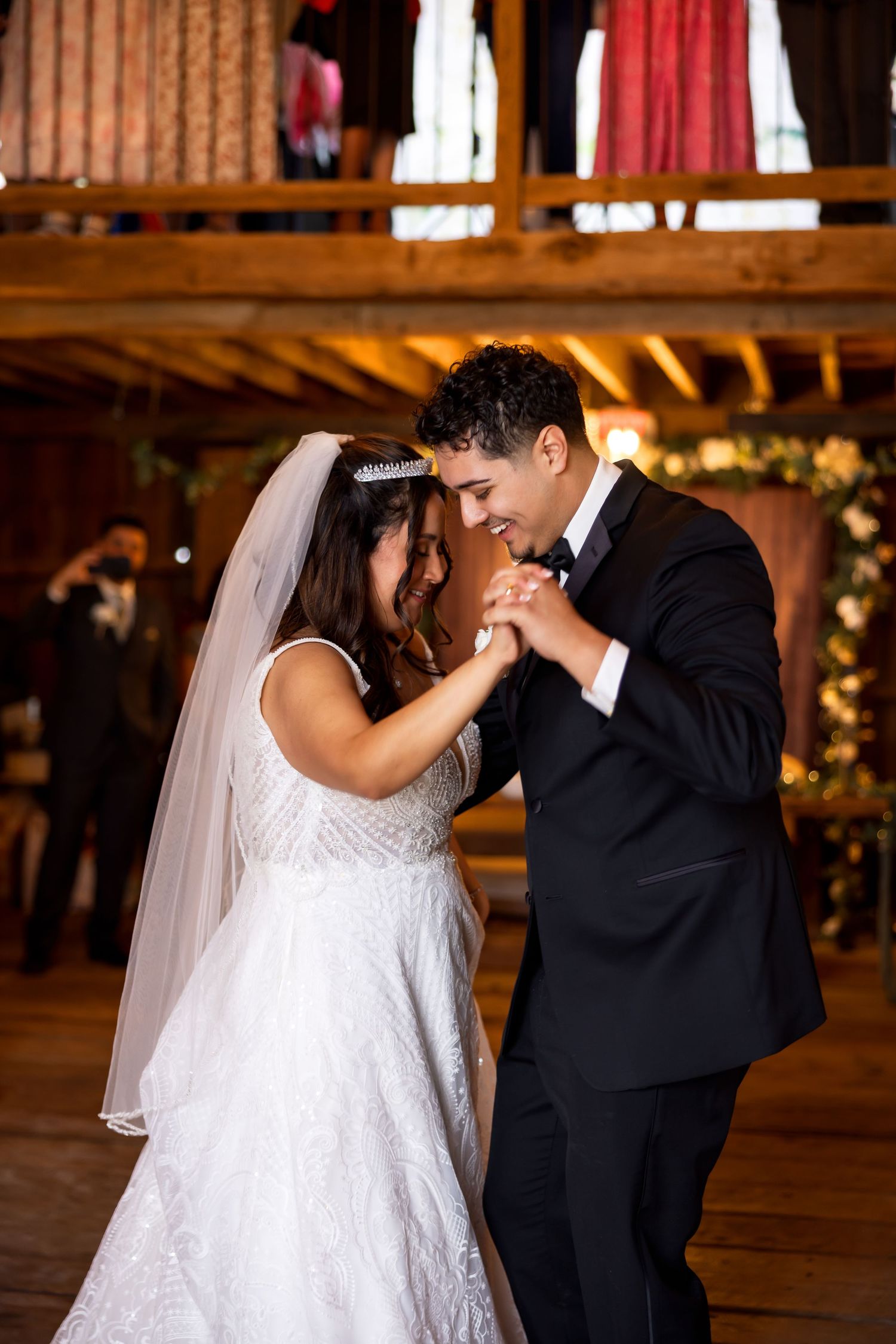 Bride and groom share their first dance in a rustic barn venue, smiling intimately while holding hands.
