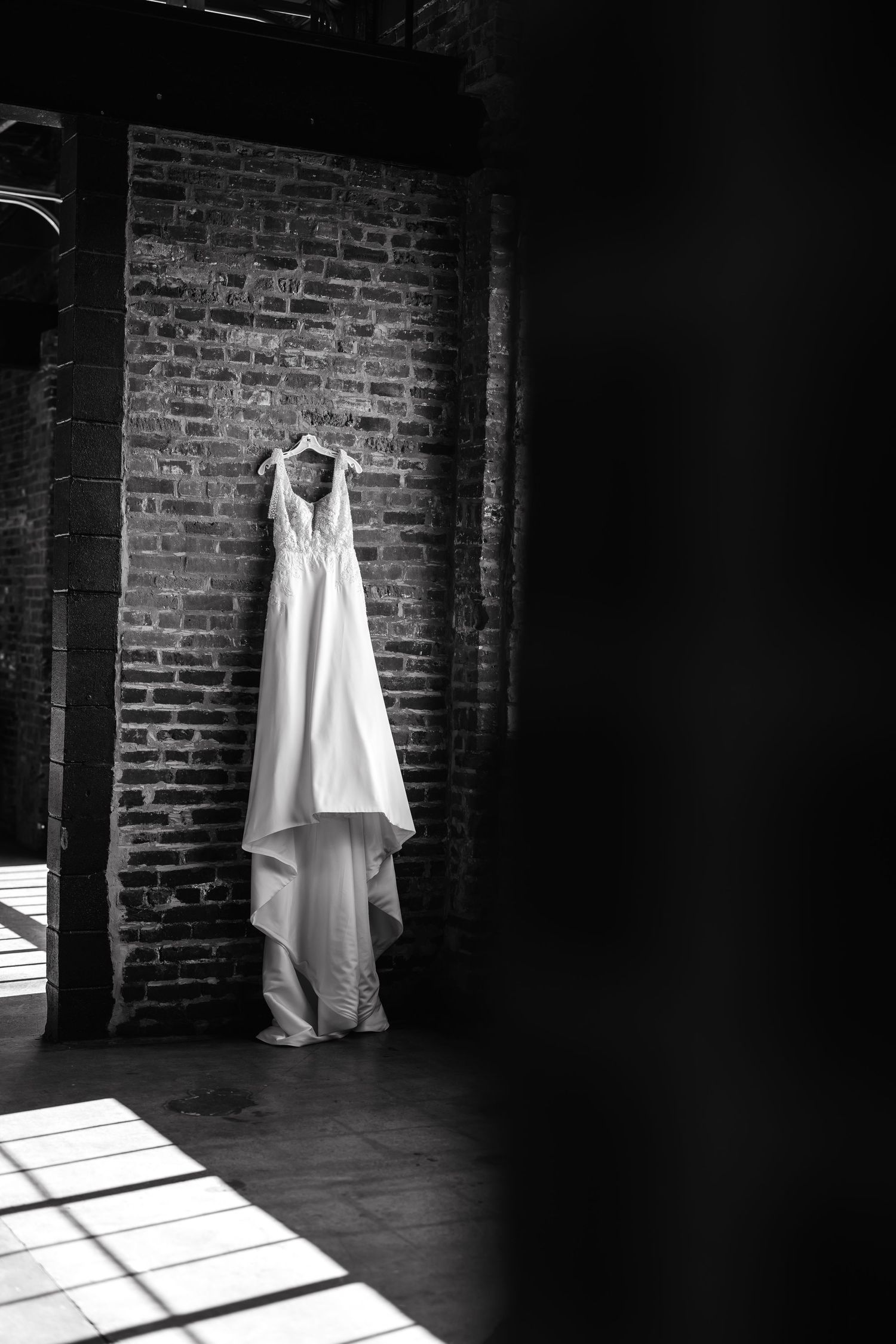 A white wedding dress hangs elegantly against a brick wall in a moody black and white composition with dramatic lighting.