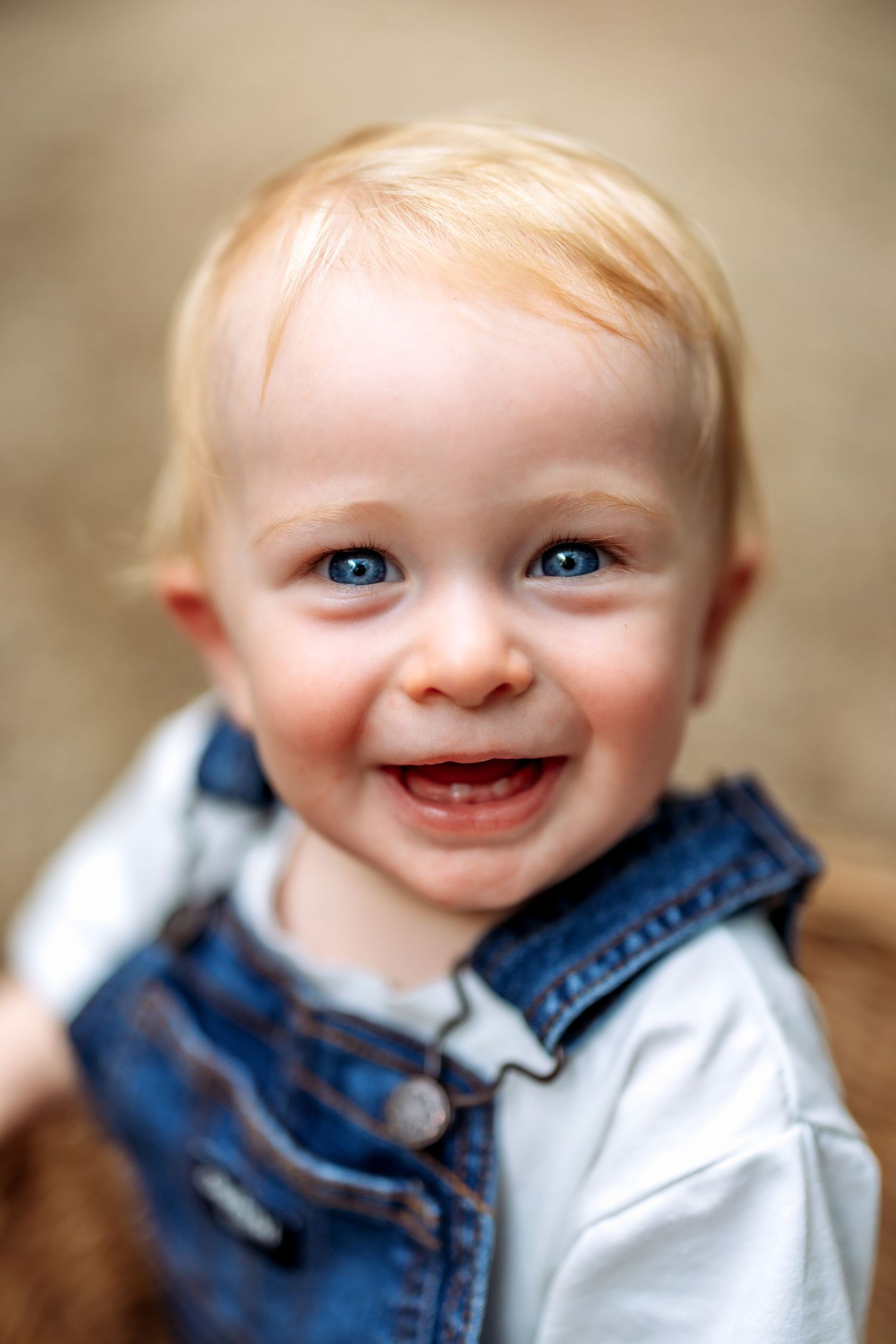 Smiling baby with blue eyes and blonde hair wearing a denim vest over a white shirt.