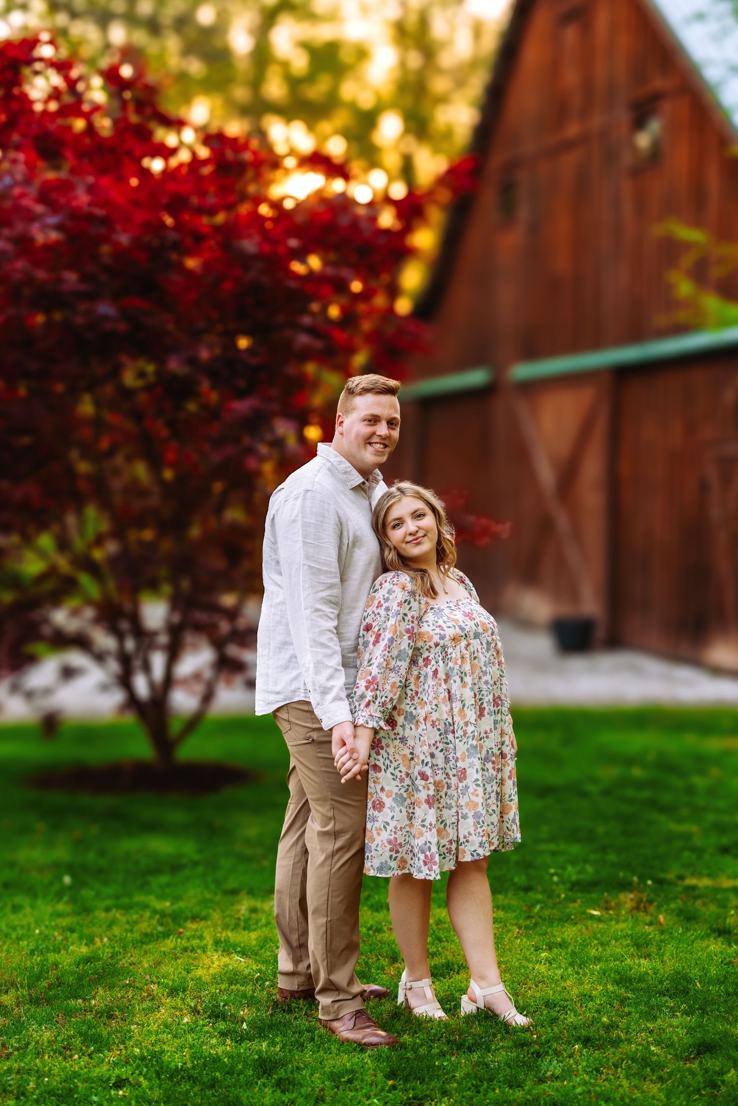 Couple standing in front of red maple tree and rustic barn during autumn.