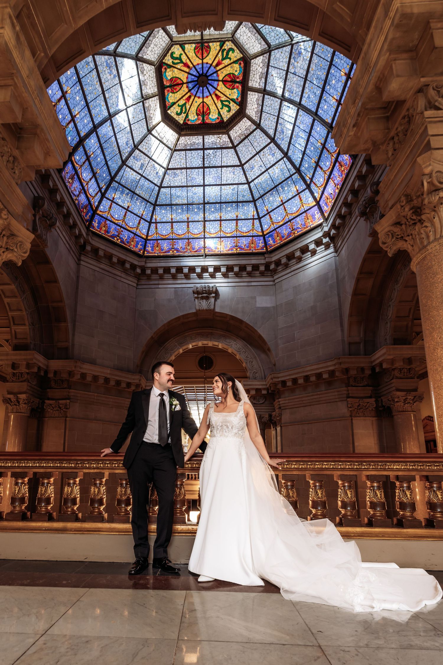 Wedding couple stands beneath a stunning octagonal stained glass dome and arched architecture in an ornate historic building.