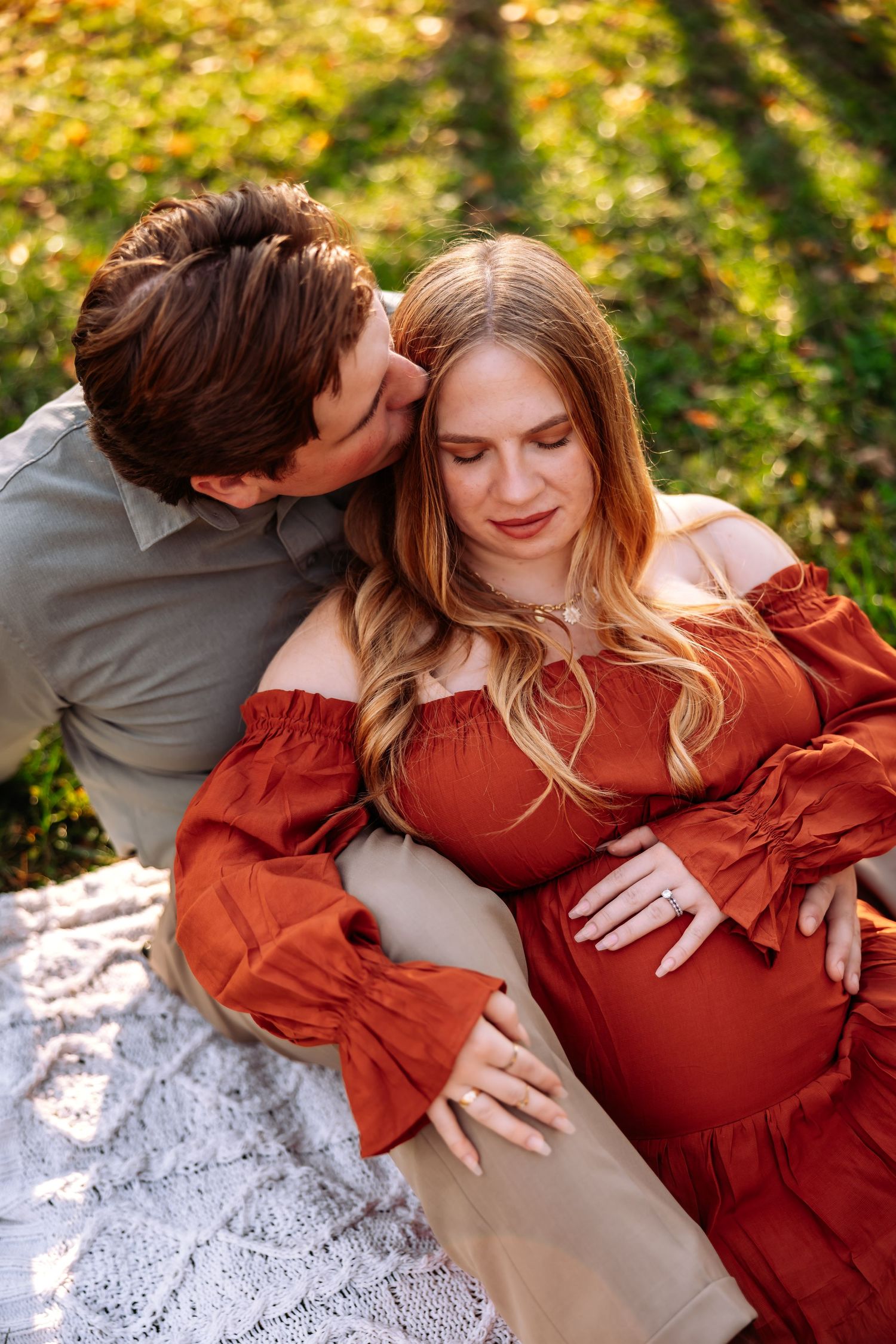 Intimate maternity photo of couple sitting on blanket outdoors with autumn foliage in background.