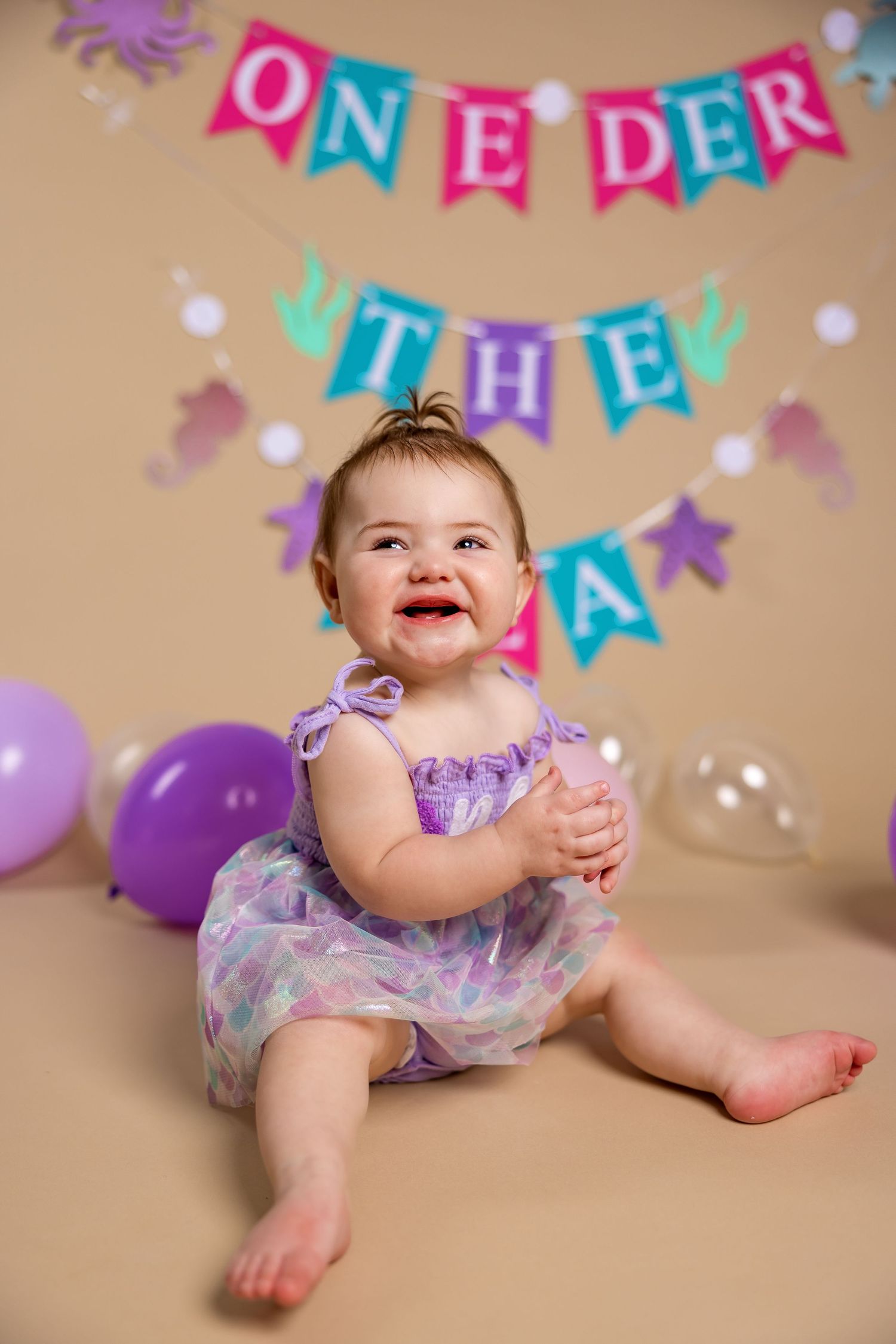 First birthday photo setup with colorful banner, purple and clear balloons, and baby in sparkly purple tulle dress.