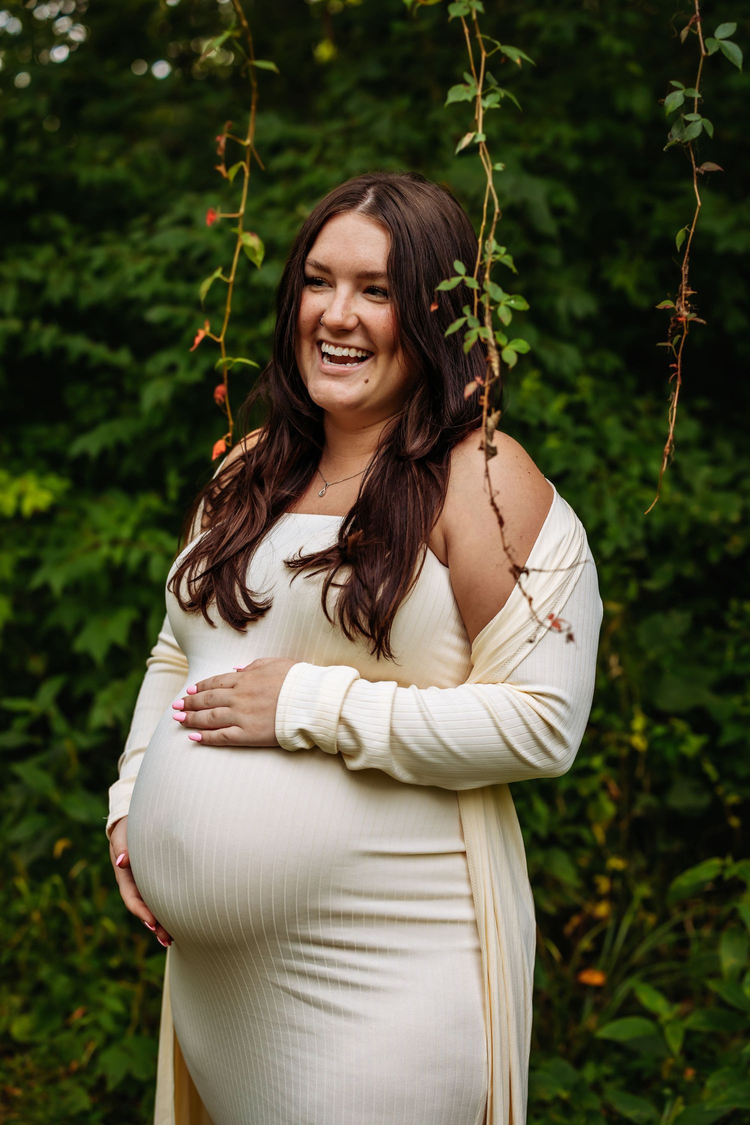 Expectant mother in white dress and cardigan stands outdoors smiling while holding her baby bump in a natural setting.