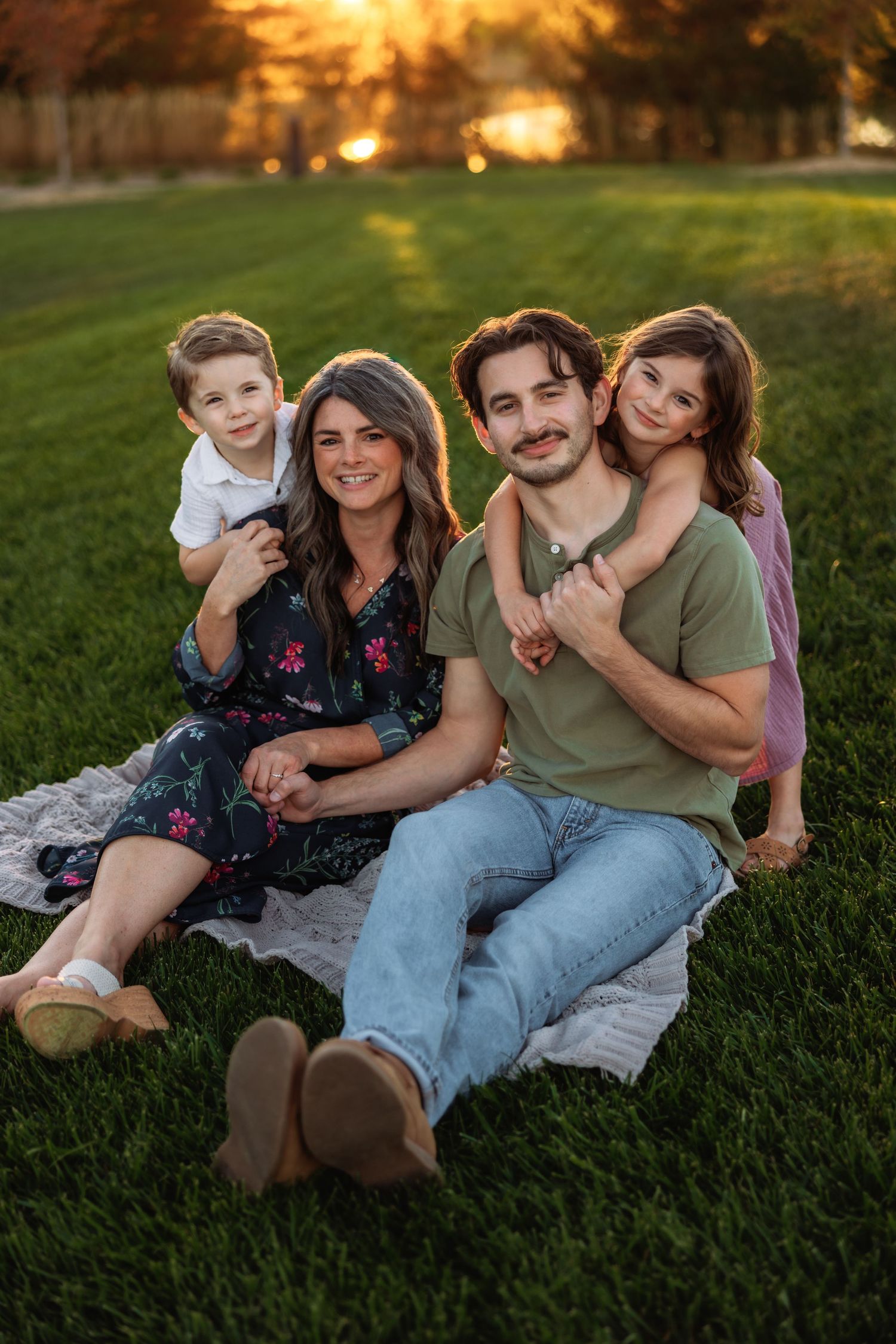A family relaxes together on green grass during golden hour sunset creating a warm outdoor portrait scene.