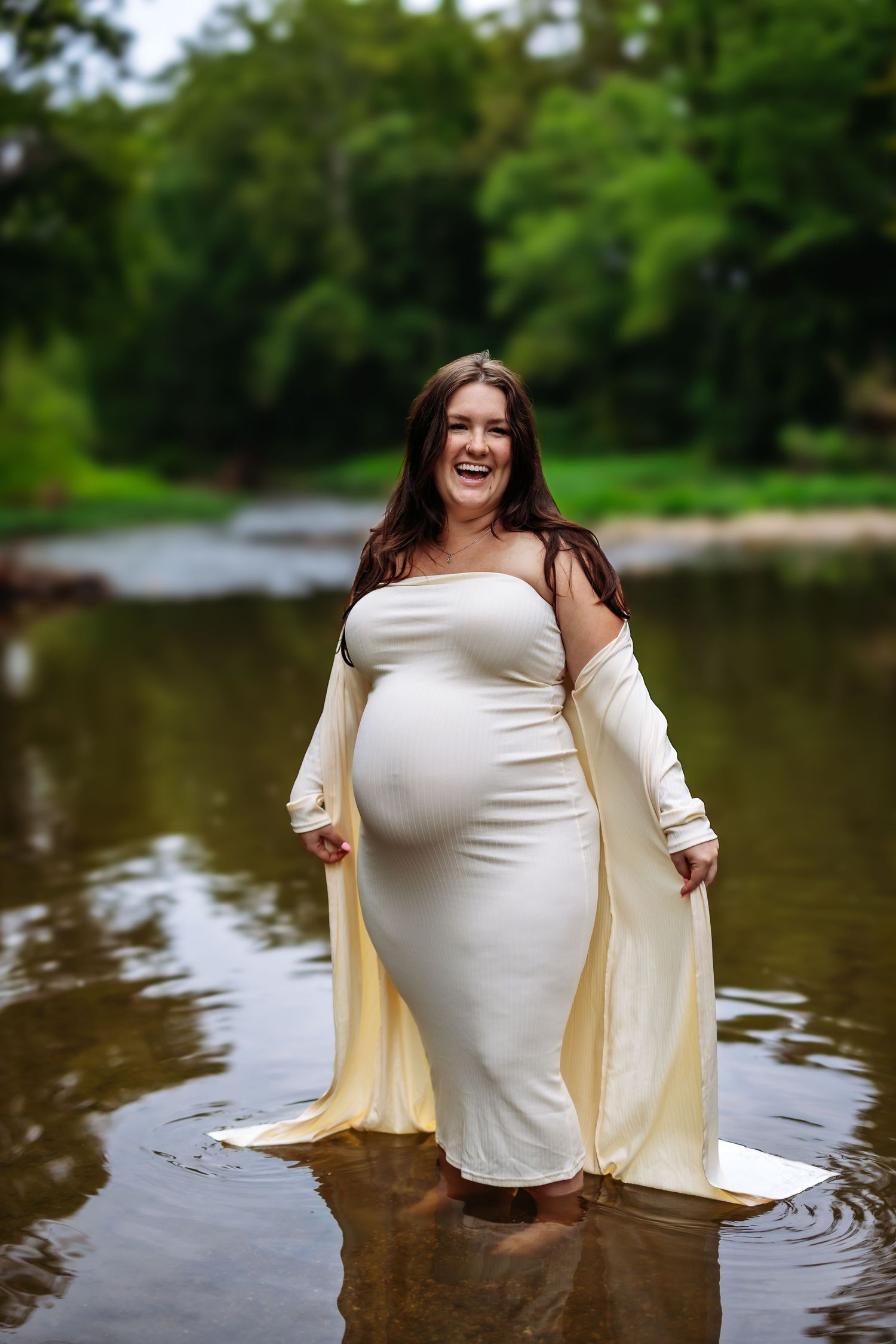 A person wearing a white dress and beige cardigan poses playfully in a lake with trees in the background.