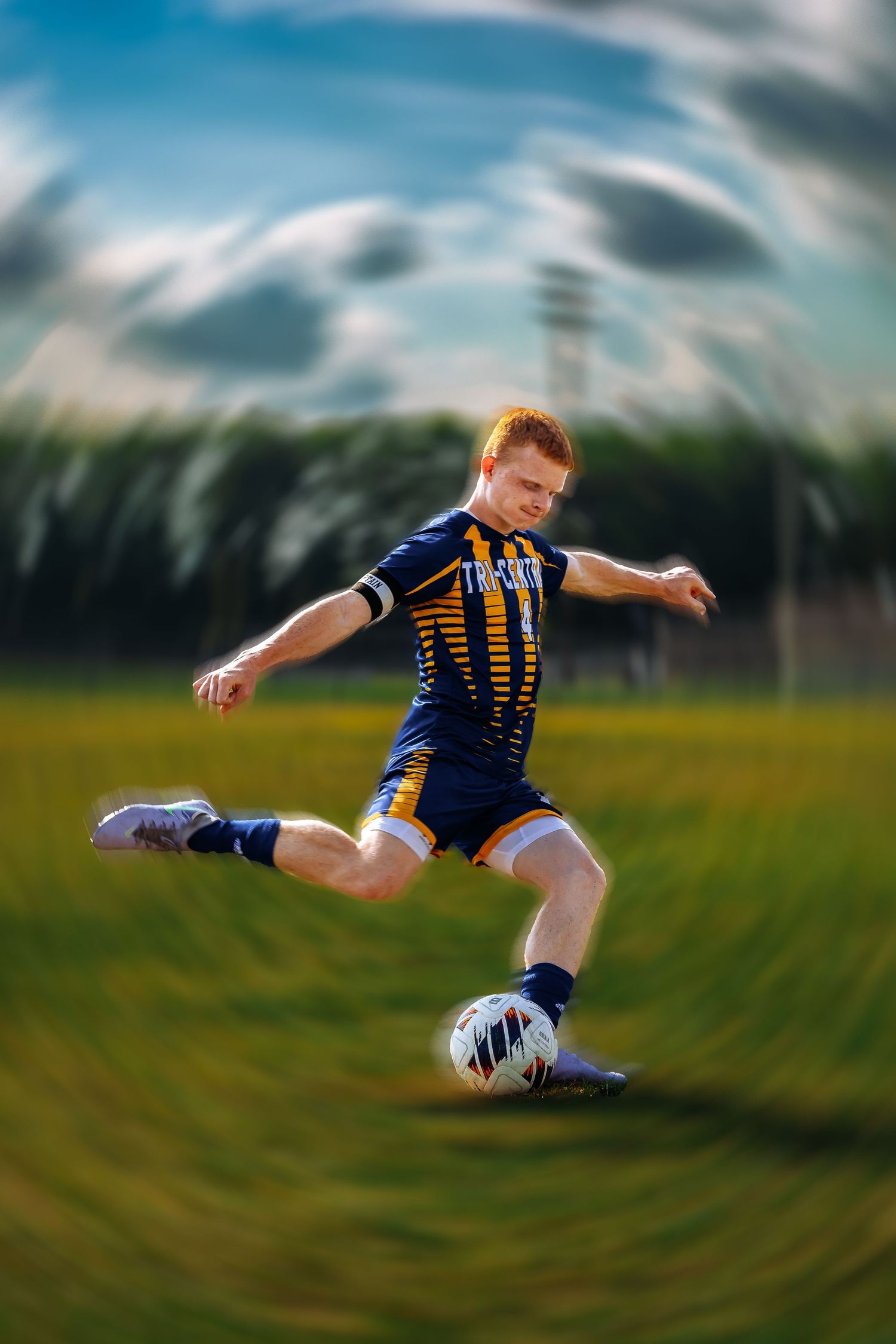 Soccer player in blue and yellow striped uniform kicking ball on grass field with motion blur effect in background.