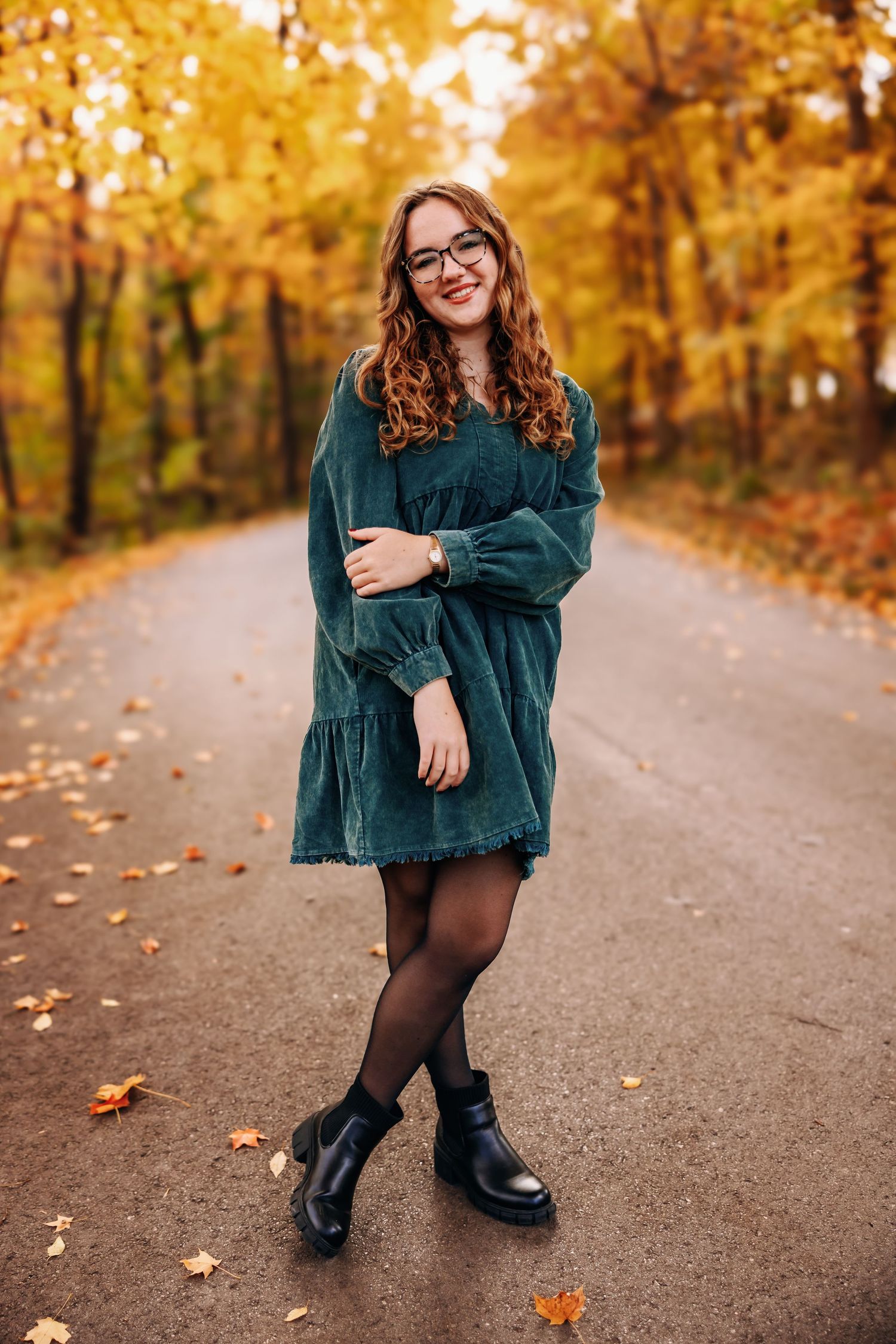 A person in a teal corduroy jacket and black boots poses on a path surrounded by golden autumn foliage.