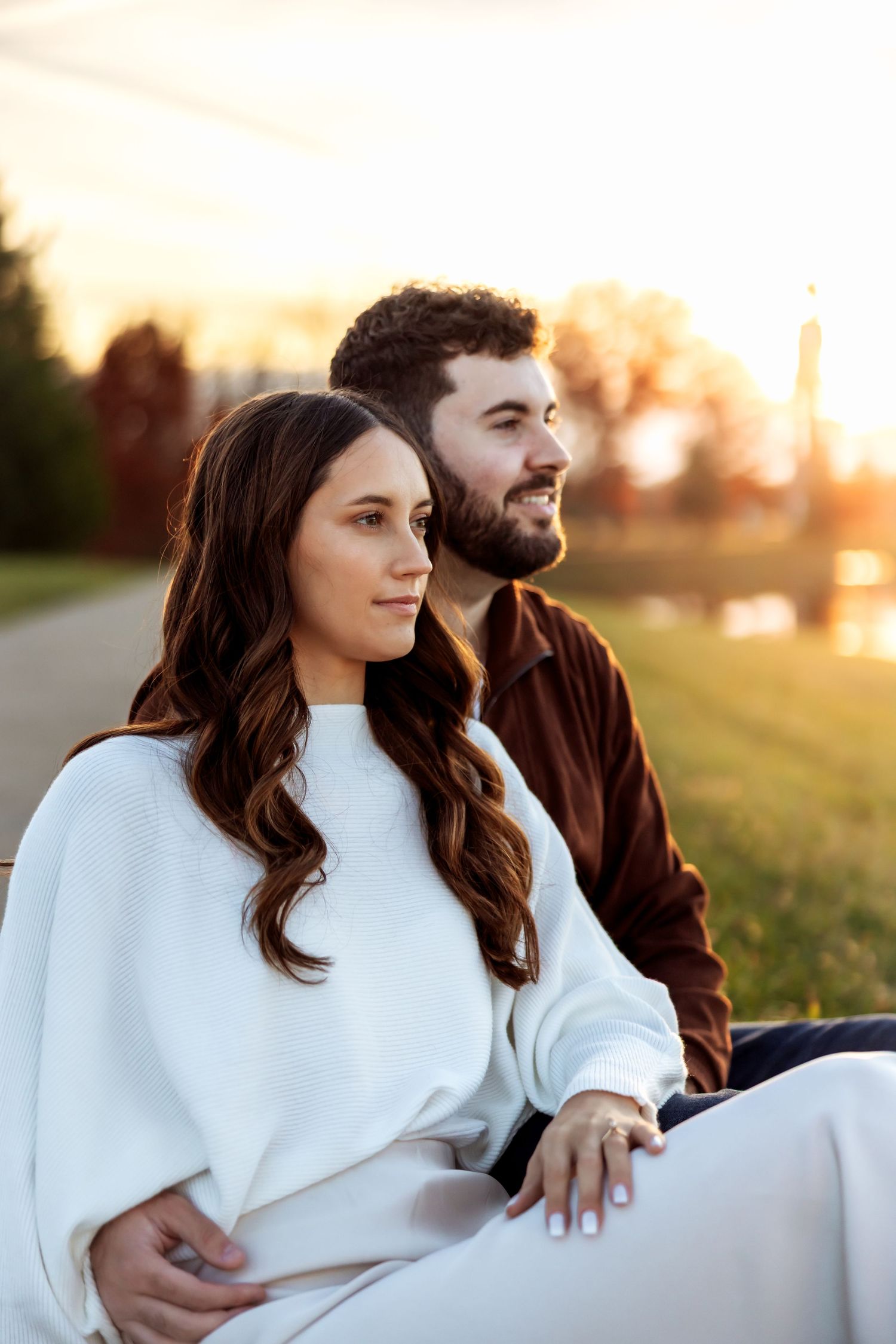 A couple sits together outdoors at sunset wrapped in a cozy white blanket in a grassy field.