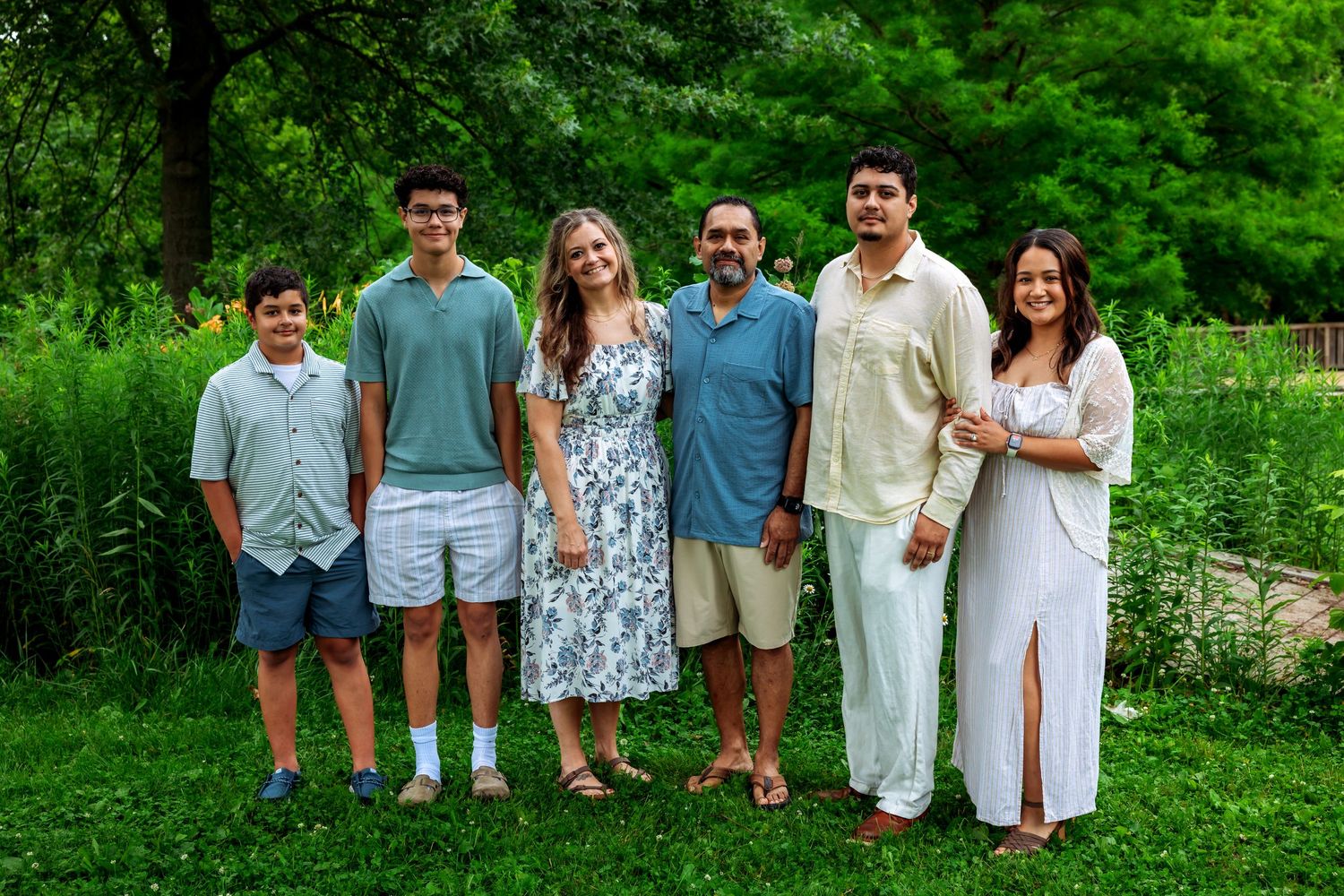 A happy family of six poses together outdoors in a lush green park setting for a summer portrait.