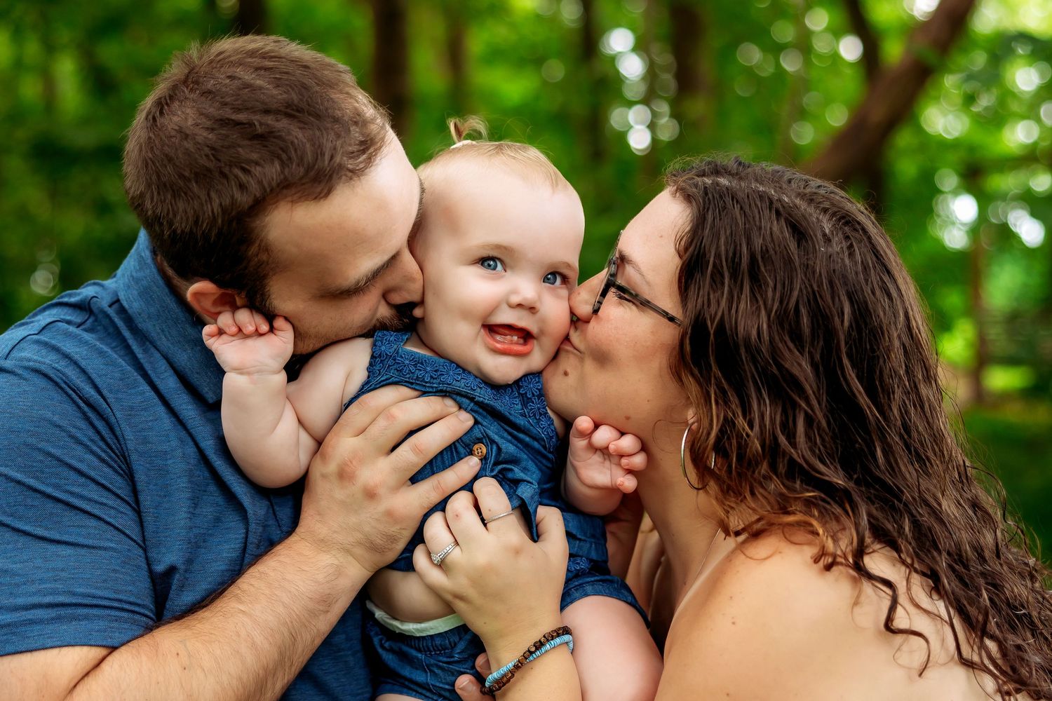 Two parents lovingly kiss their smiling baby outdoors, surrounded by lush green trees in a heartwarming family portrait.