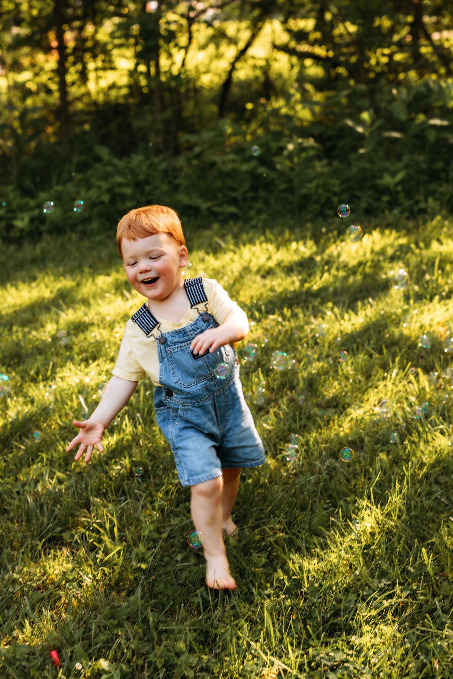 Joyful toddler in denim overalls running barefoot through sunny grass with bubbles floating around.
