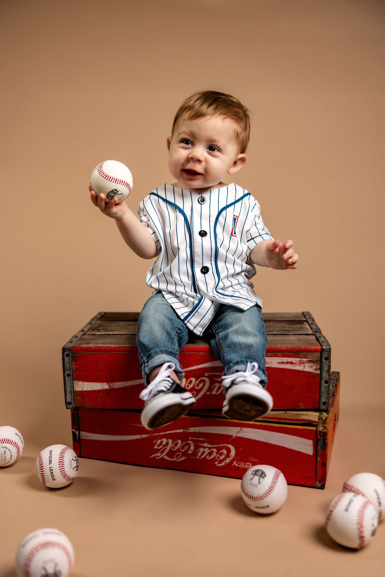 A toddler in a pinstriped baseball jersey sits on a vintage Coca-Cola crate surrounded by baseballs.