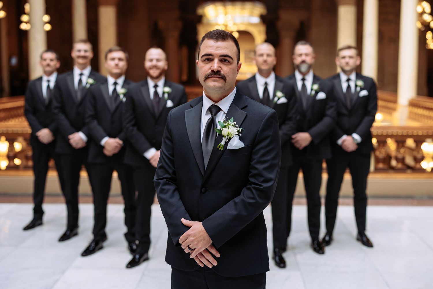 A groom stands in front with groomsmen lined up behind him in black suits at an elegant venue interior.
