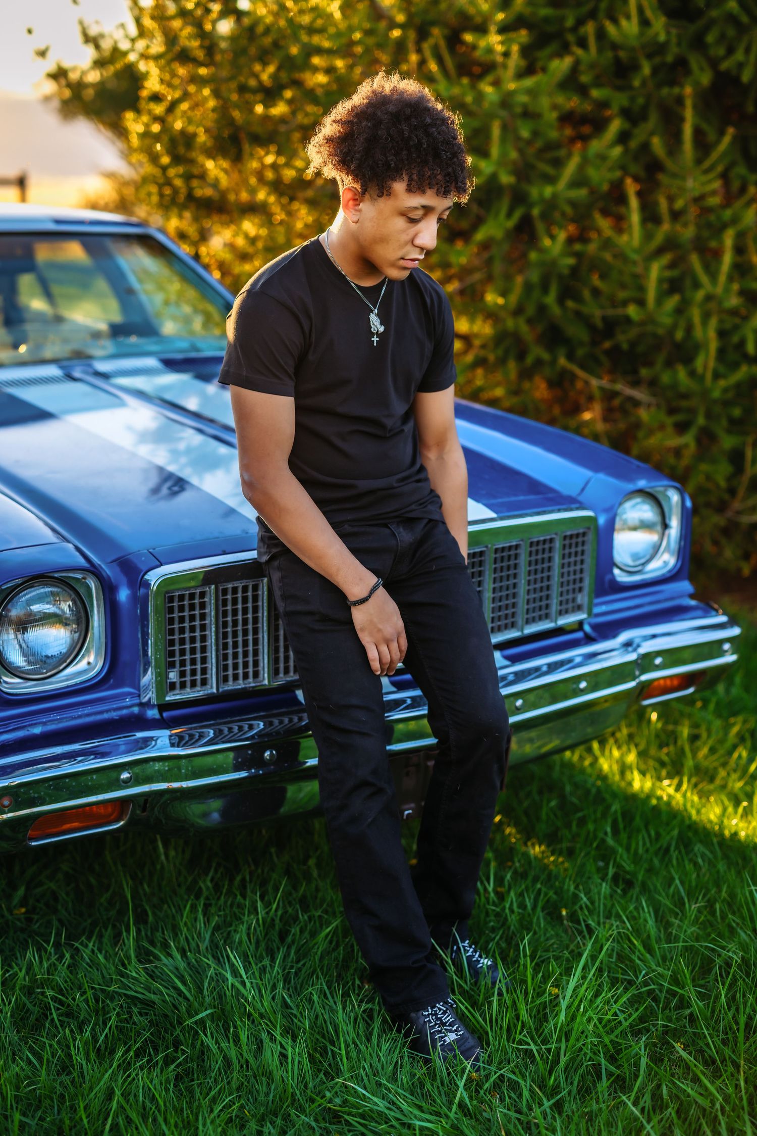 Person in black outfit poses against vintage blue car during golden hour.