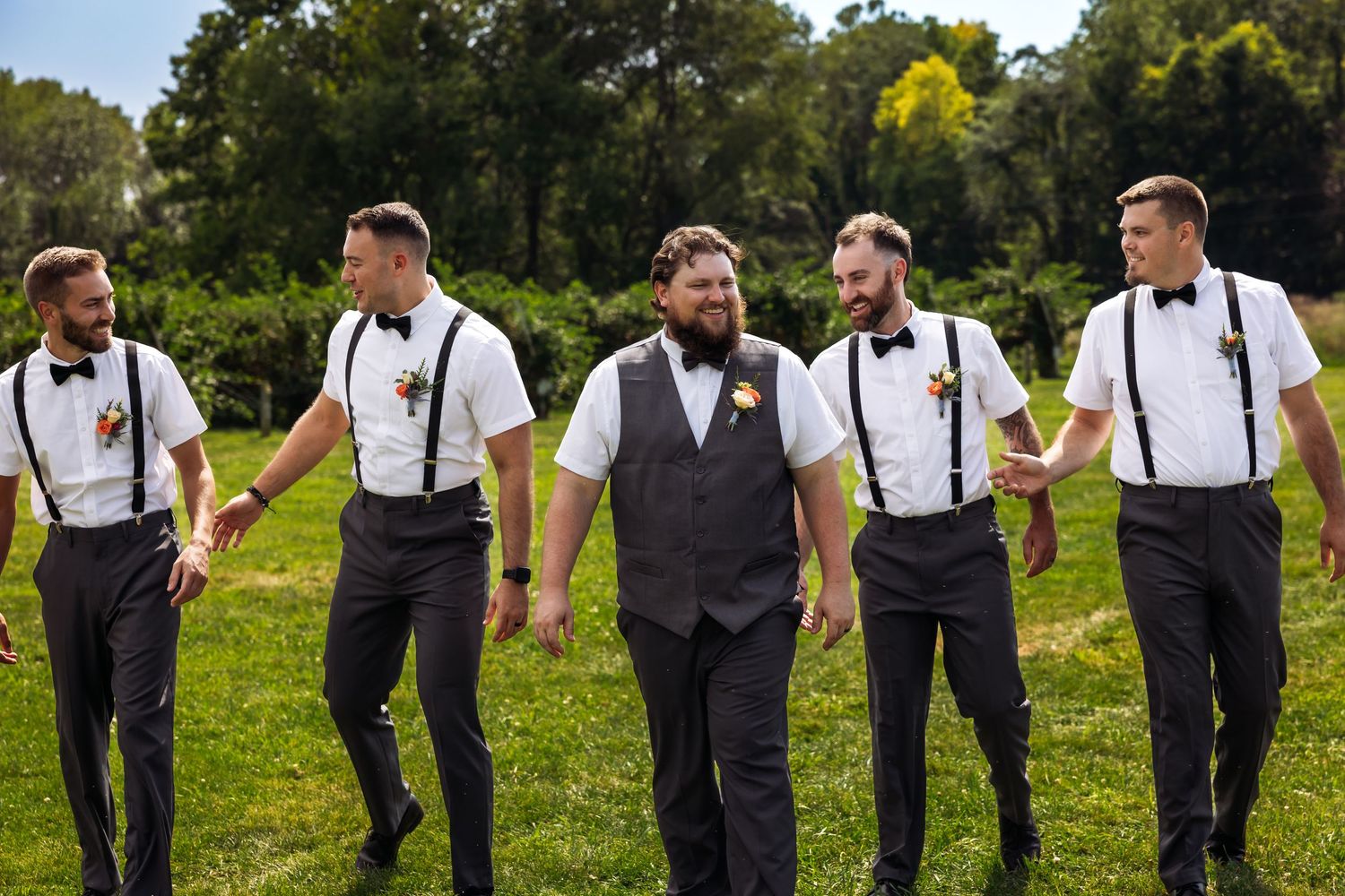 Groomsmen in matching black suspenders and white shirts walk together on green grass during outdoor wedding celebration.
