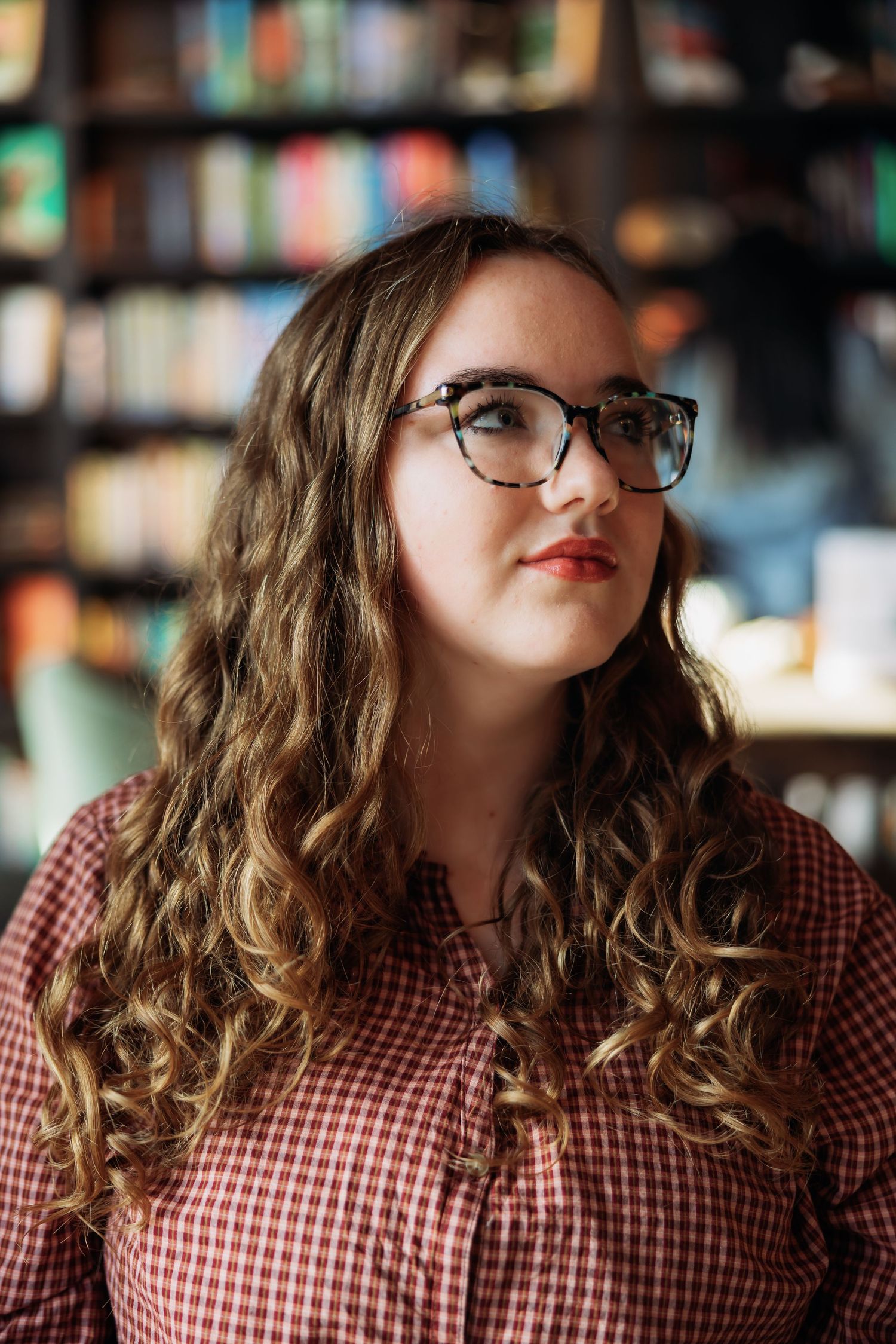 Person with curly hair and glasses wearing a red and white checkered shirt in a library setting.
