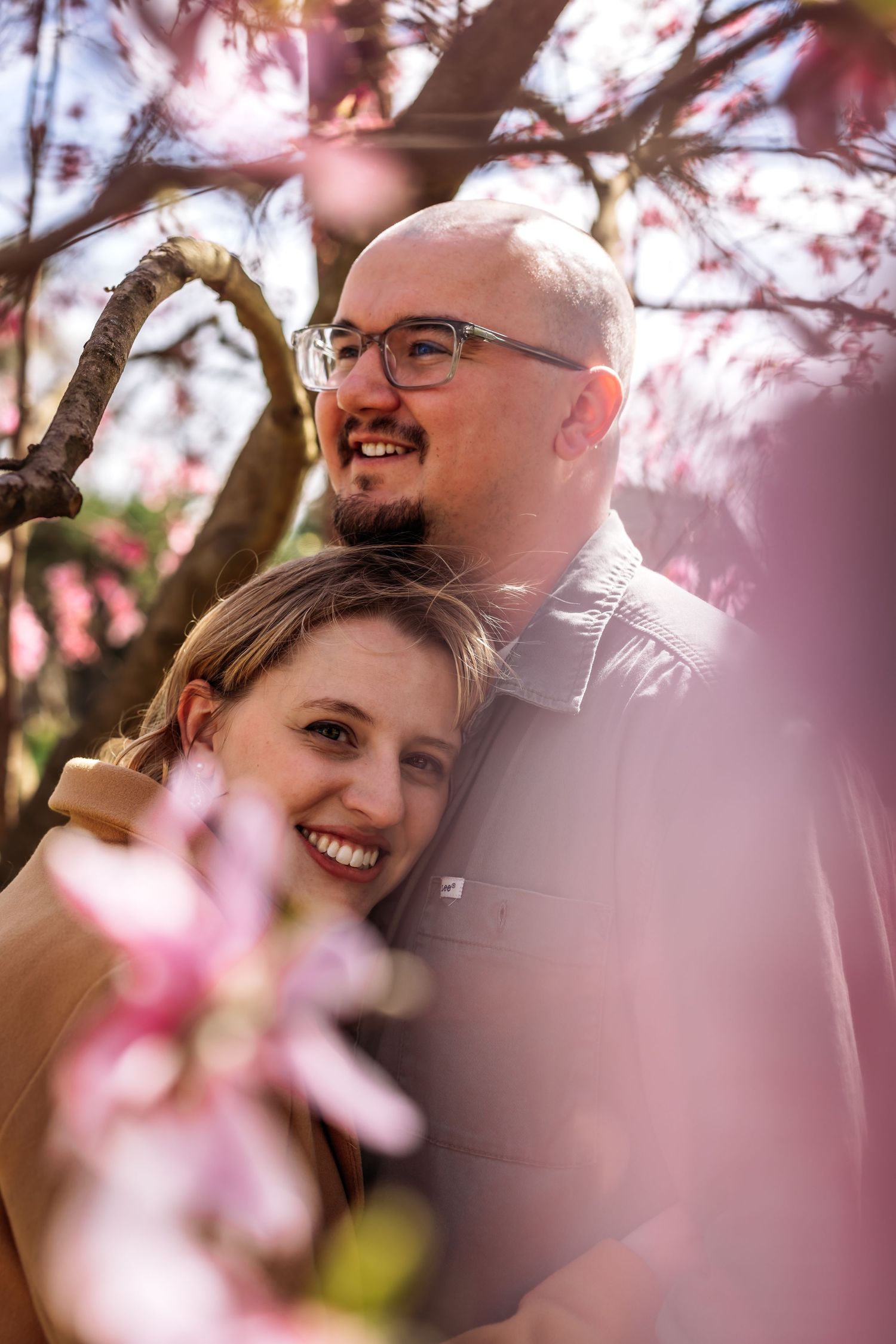 Couple smiling under blooming cherry blossom trees with soft pink petals.