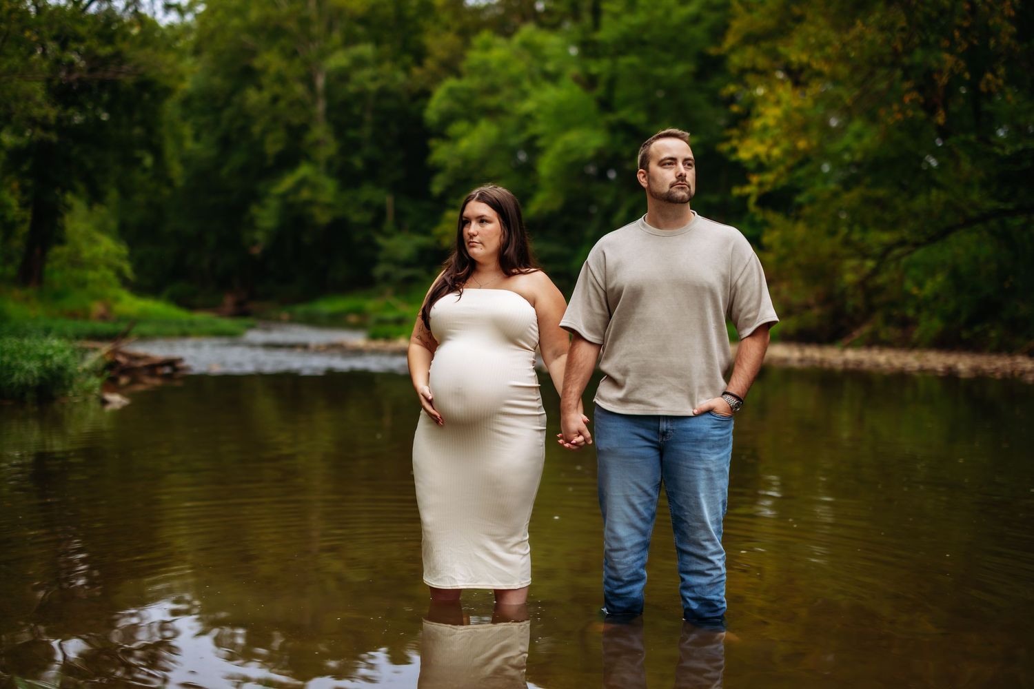 A couple stands together by a serene lake surrounded by lush greenery on a summer day.