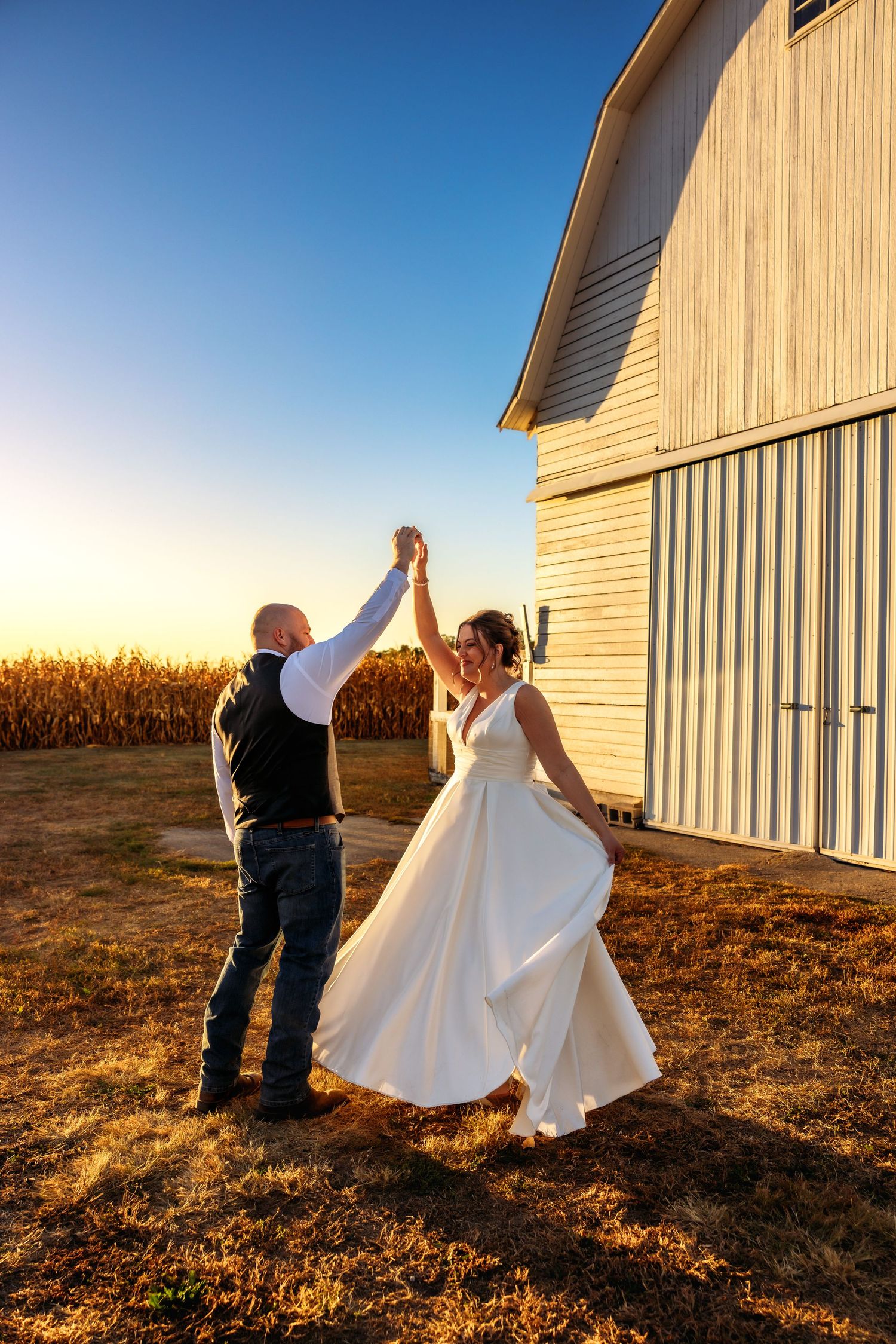A couple dances in front of rustic barn buildings at sunset with golden light casting long shadows across a field.