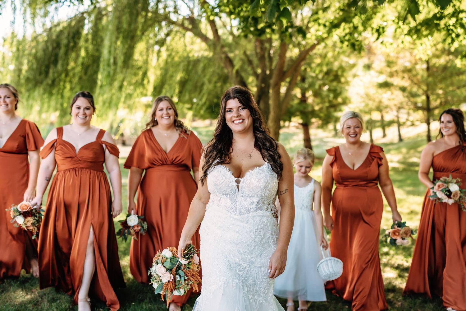 A wedding party poses outdoors wearing rust-colored bridesmaid dresses and a white wedding gown in a garden setting.