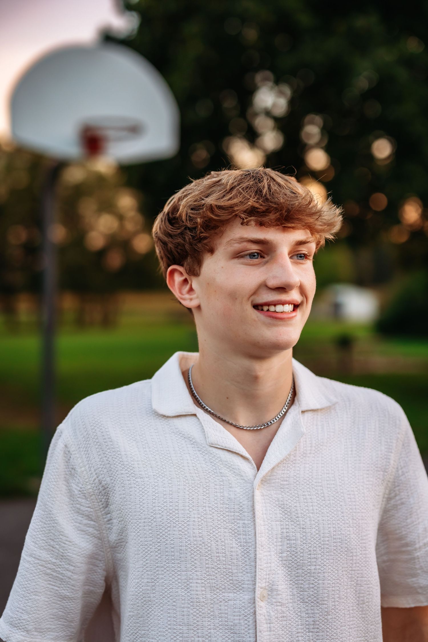 Young person in white polo shirt smiling outdoors with basketball hoop in background.