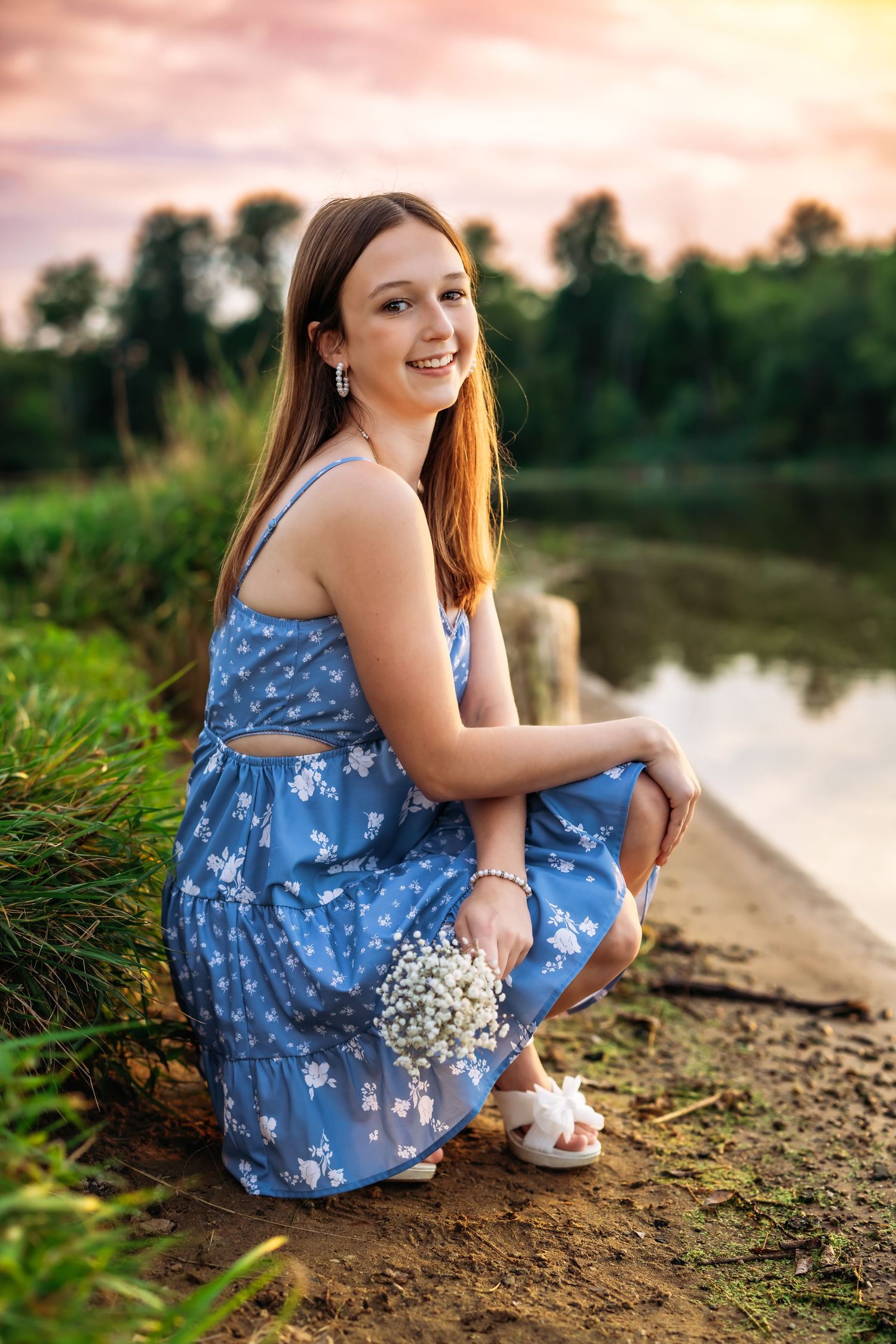 Portrait of someone in blue floral dress holding flowers by lake at sunset.