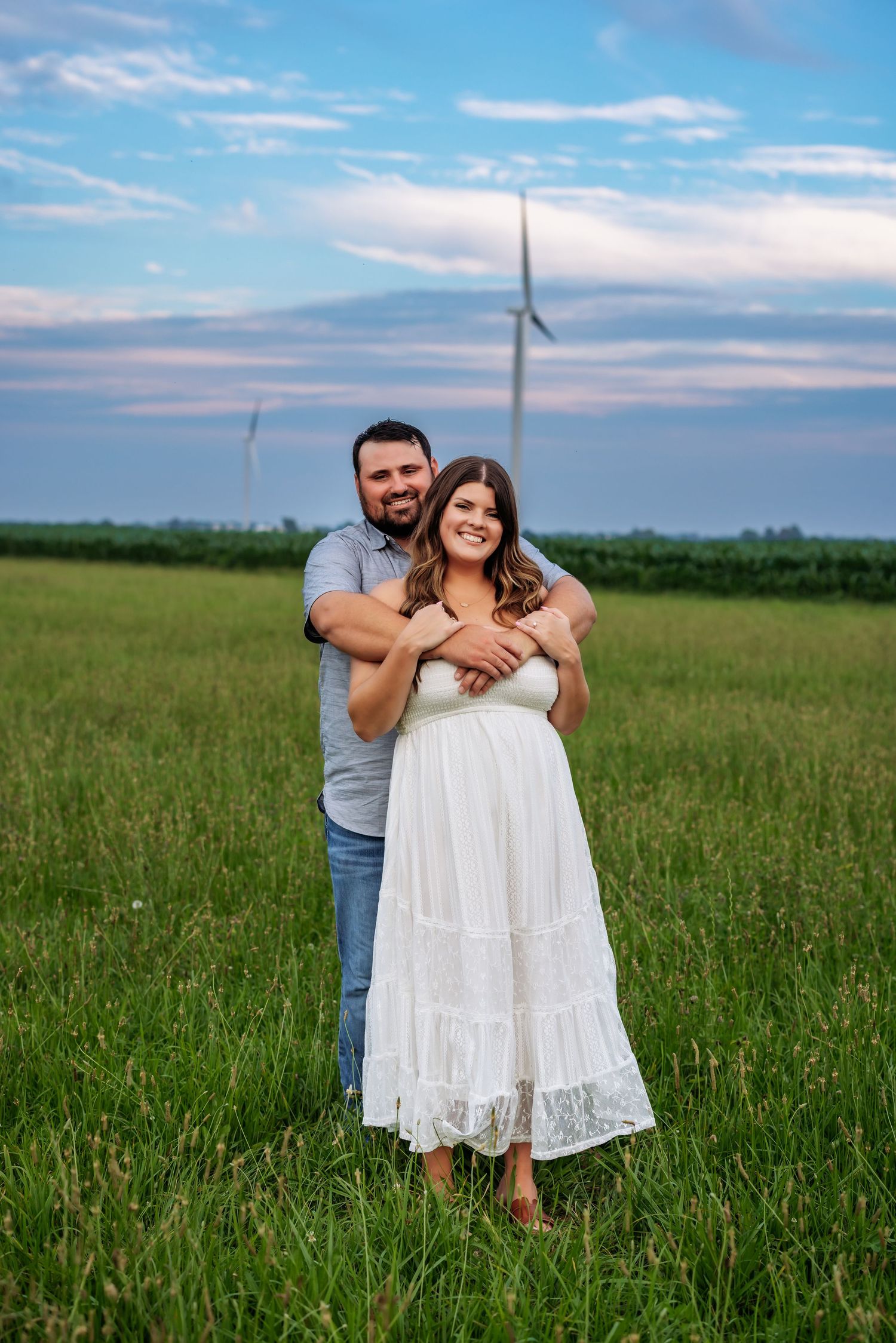 A couple embraces in a green field with wind turbines visible in the background during sunset.