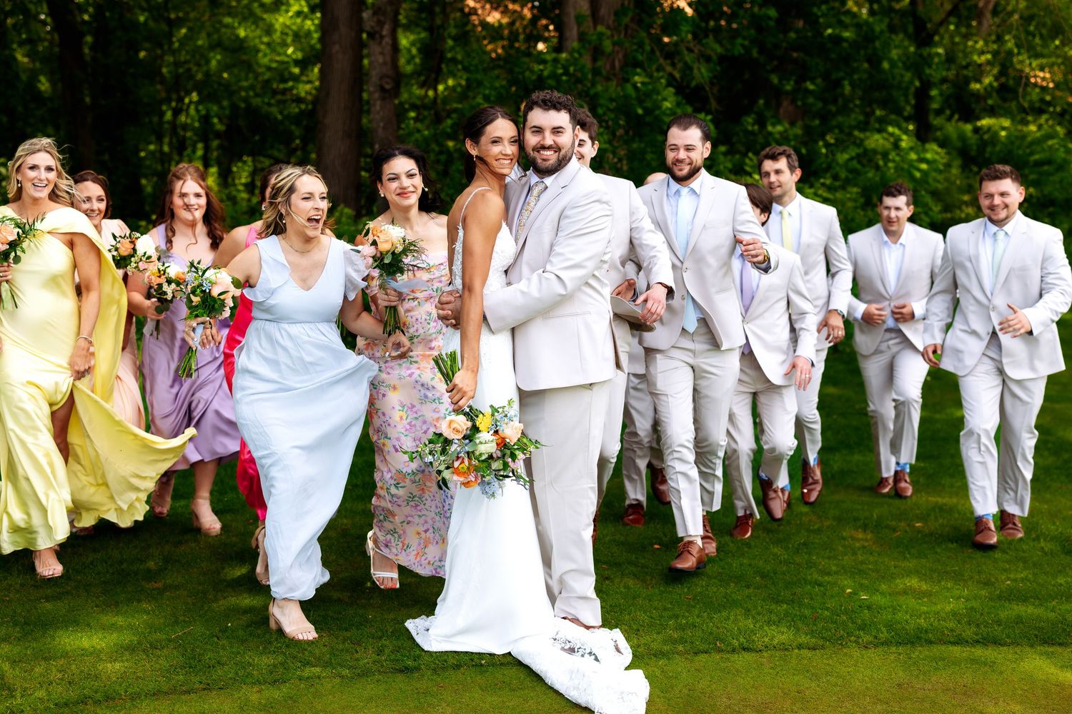 Wedding party walks outdoors, bride and groom embrace while bridesmaids in colorful dresses and groomsmen in gray suits celebrate.