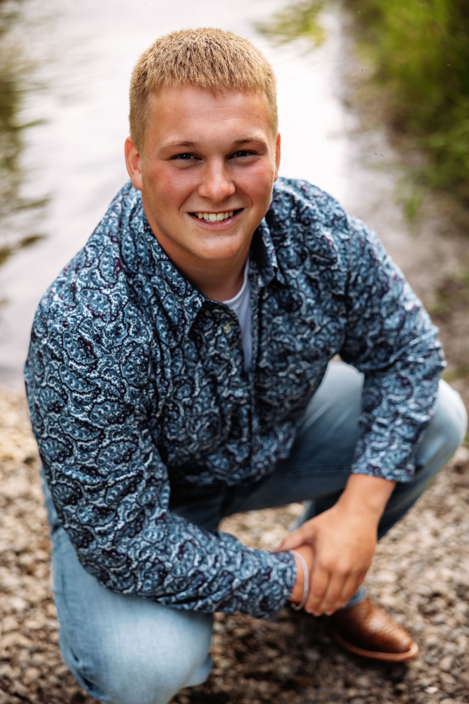 A person in a blue patterned shirt and jeans sits on a gravel path in a natural outdoor setting.