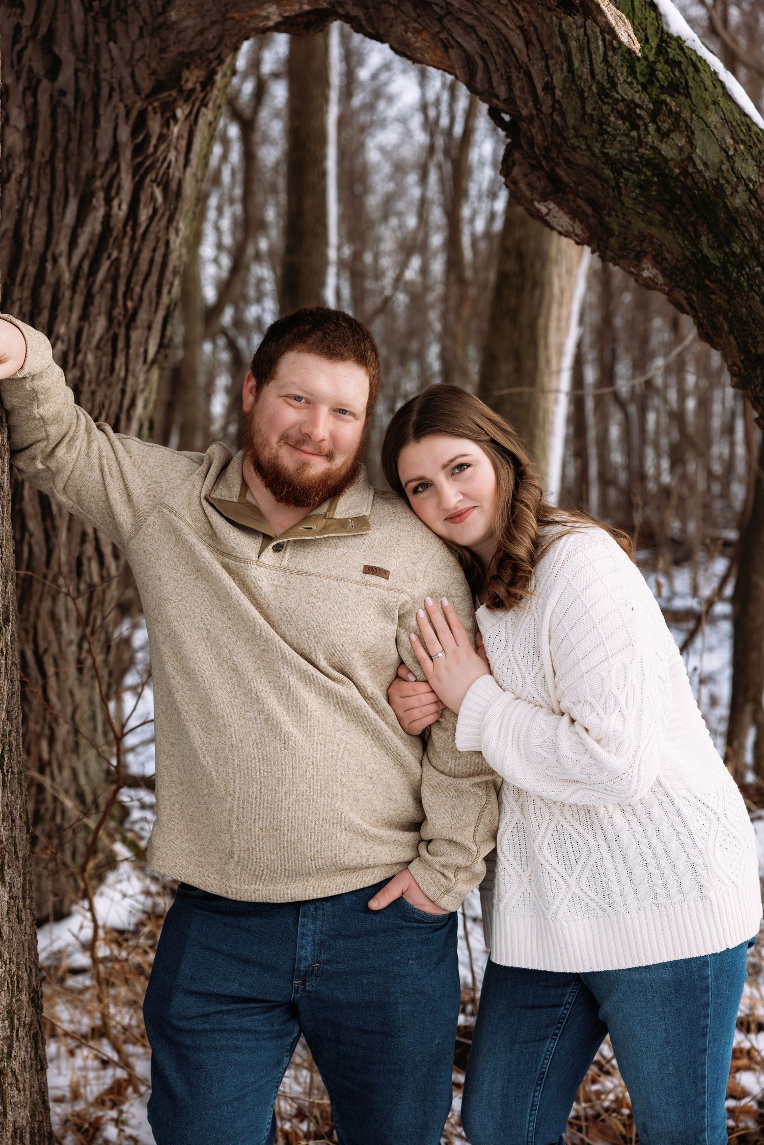 A couple poses playfully against a tree trunk in a snowy winter forest setting wearing casual sweaters and jeans.