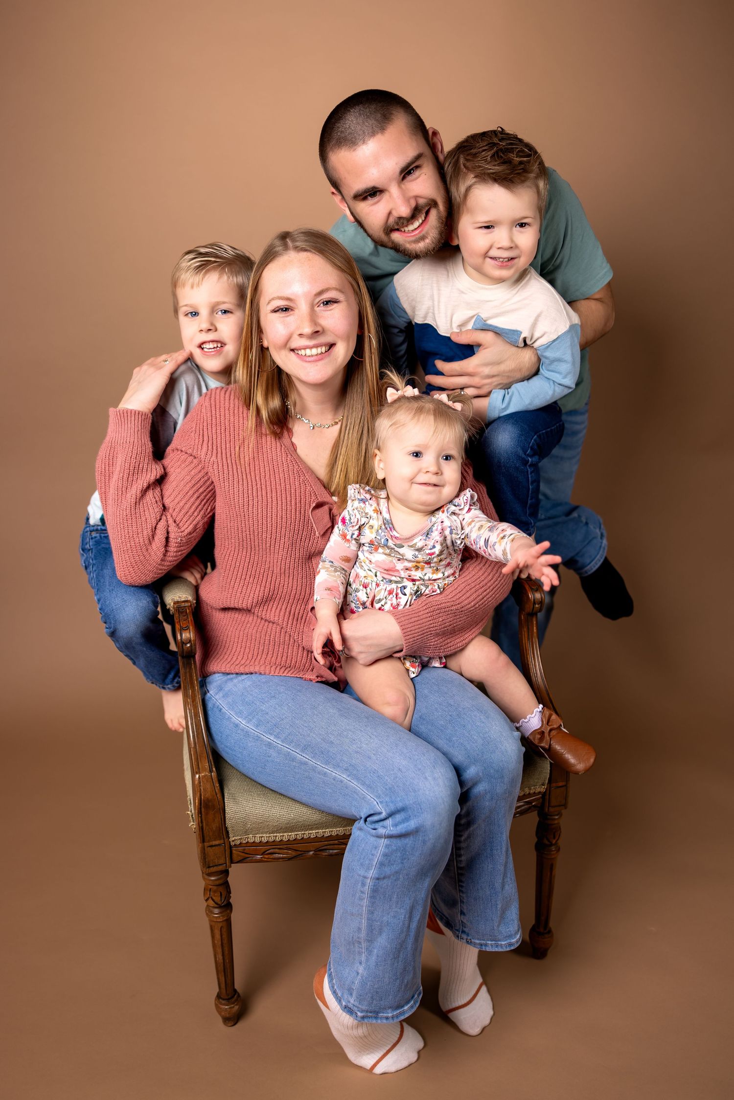 A family poses together on a wooden chair in a studio with brown backdrop showing playful expressions and movements.