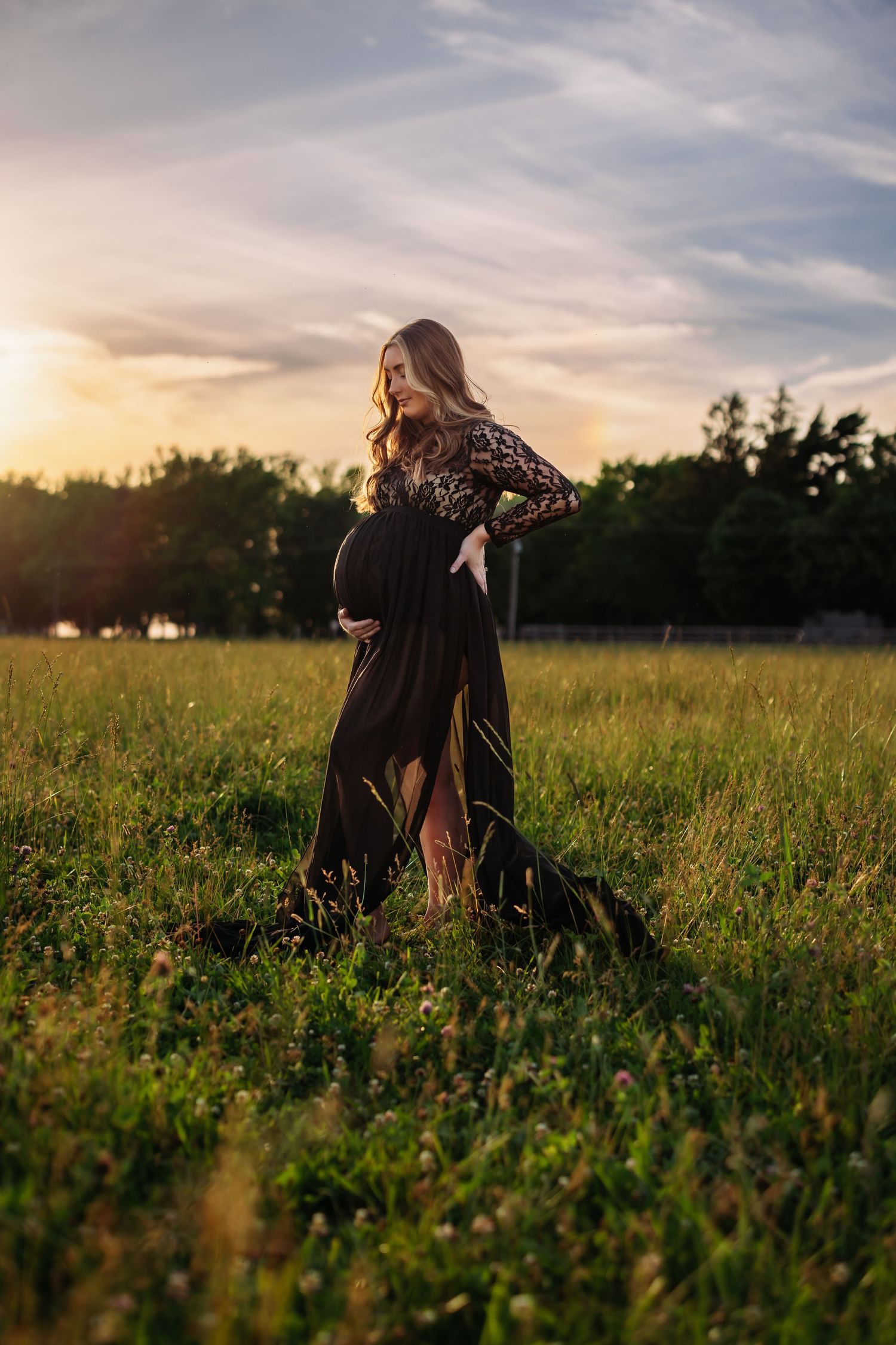 Maternity photo of woman in black lace dress during golden hour in grassy field.