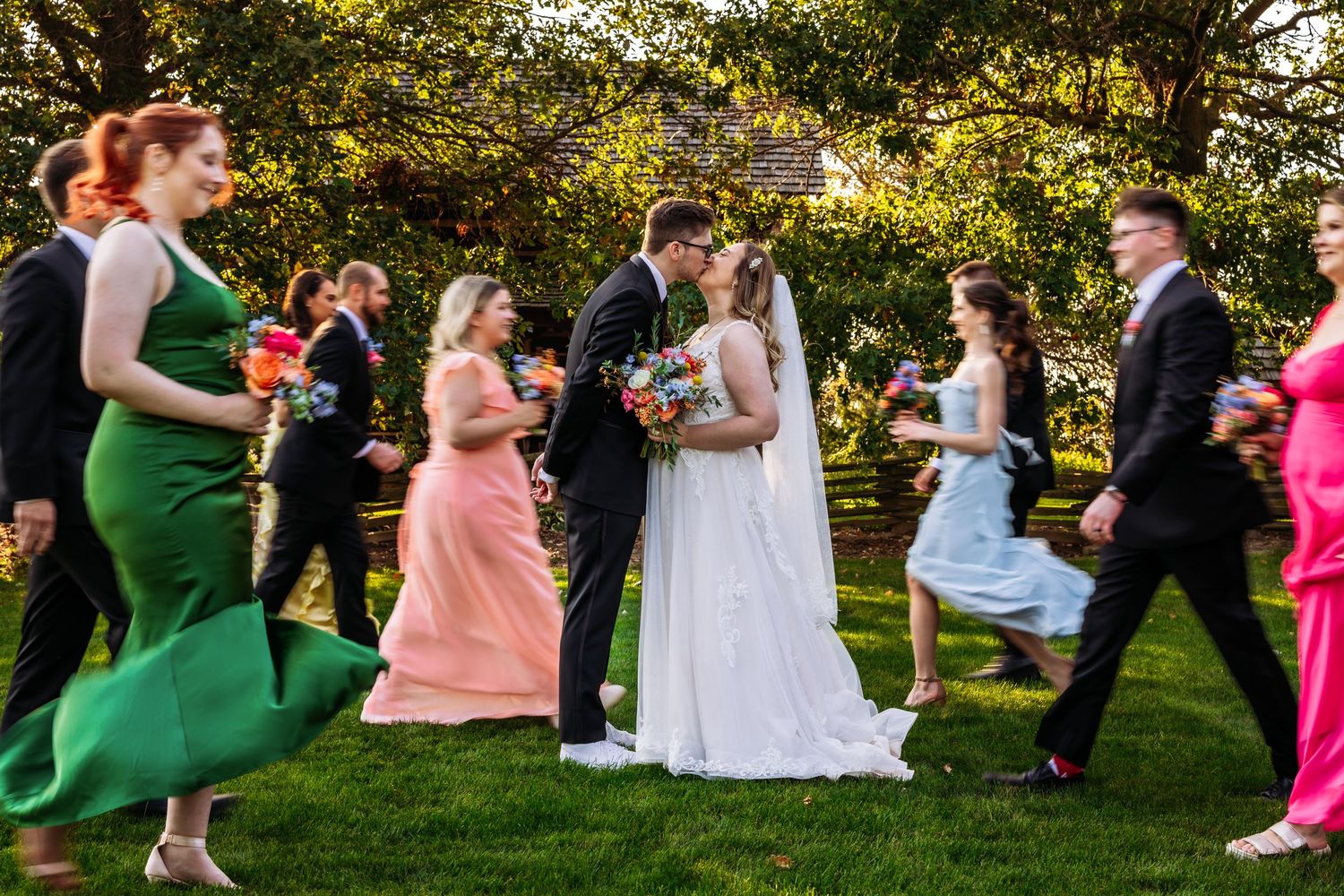 Wedding party in colorful dresses dances joyfully on a sunlit lawn.