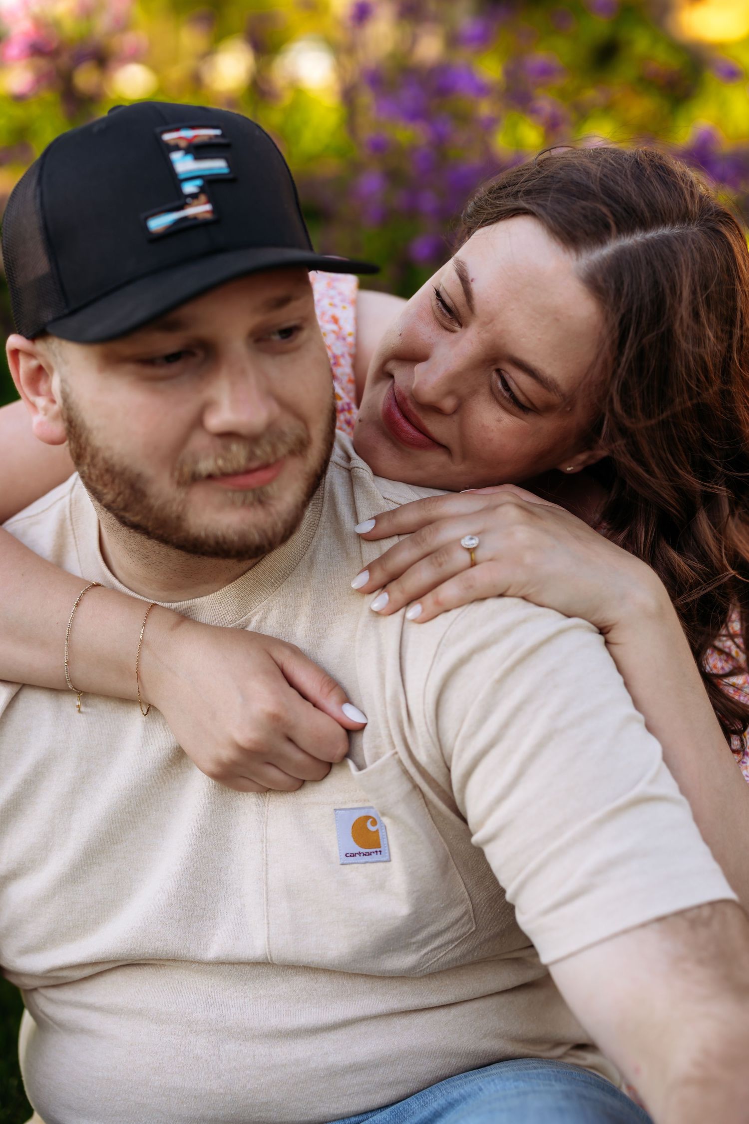 A couple embraces and shares tender moments in a garden with purple flowers blooming in the background.