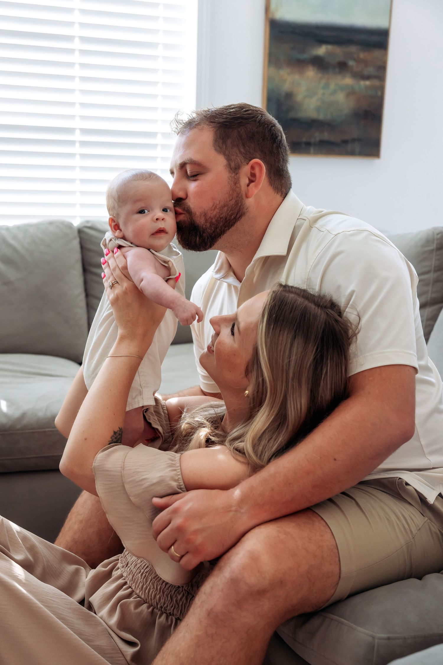 A couple sits on a couch, kissing their newborn baby in a bright living room.