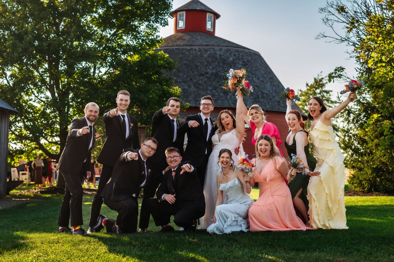 Wedding party poses playfully for a group photo in front of a round barn structure.